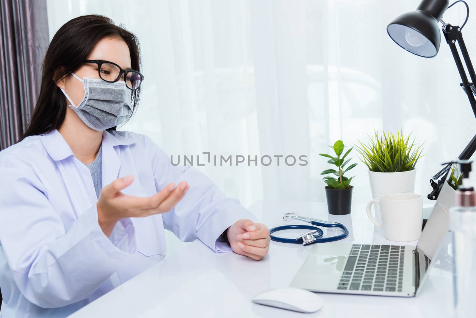 Asian young beautiful woman doctor wear face mask protective video call conference online raise hand explain to patients on laptop computer quarantines disease coronavirus or COVID-19 at hospital