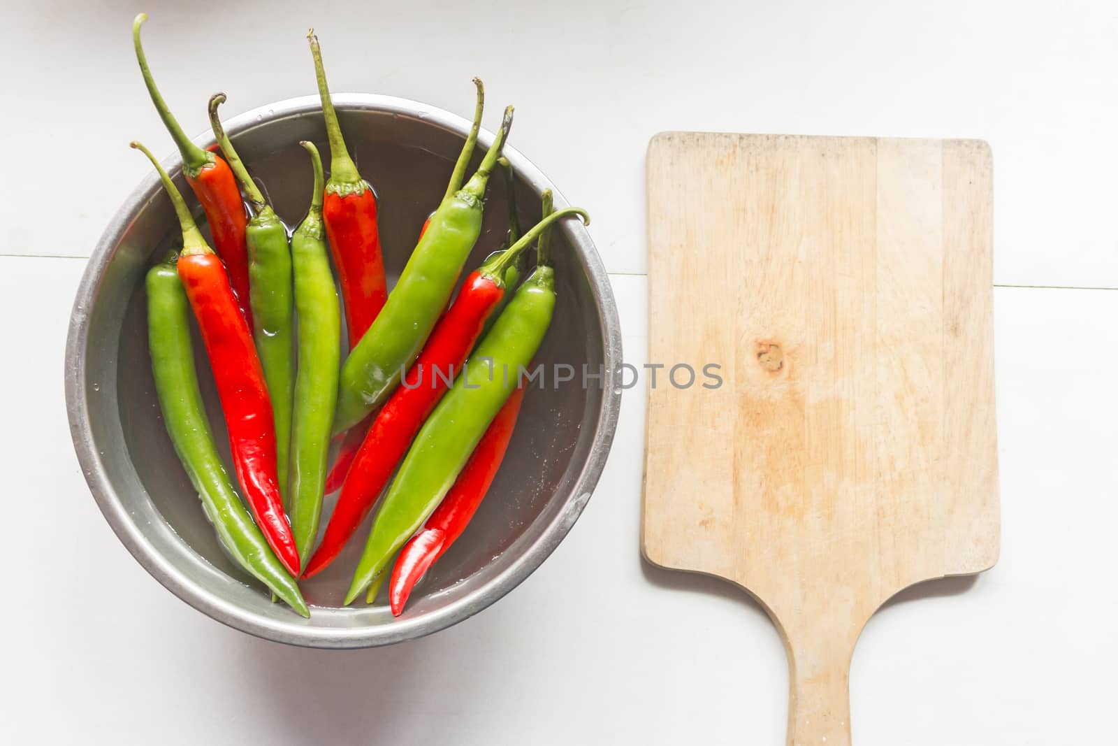 Red and green chilli pepper in water on bowl of steel with cutting board, top view