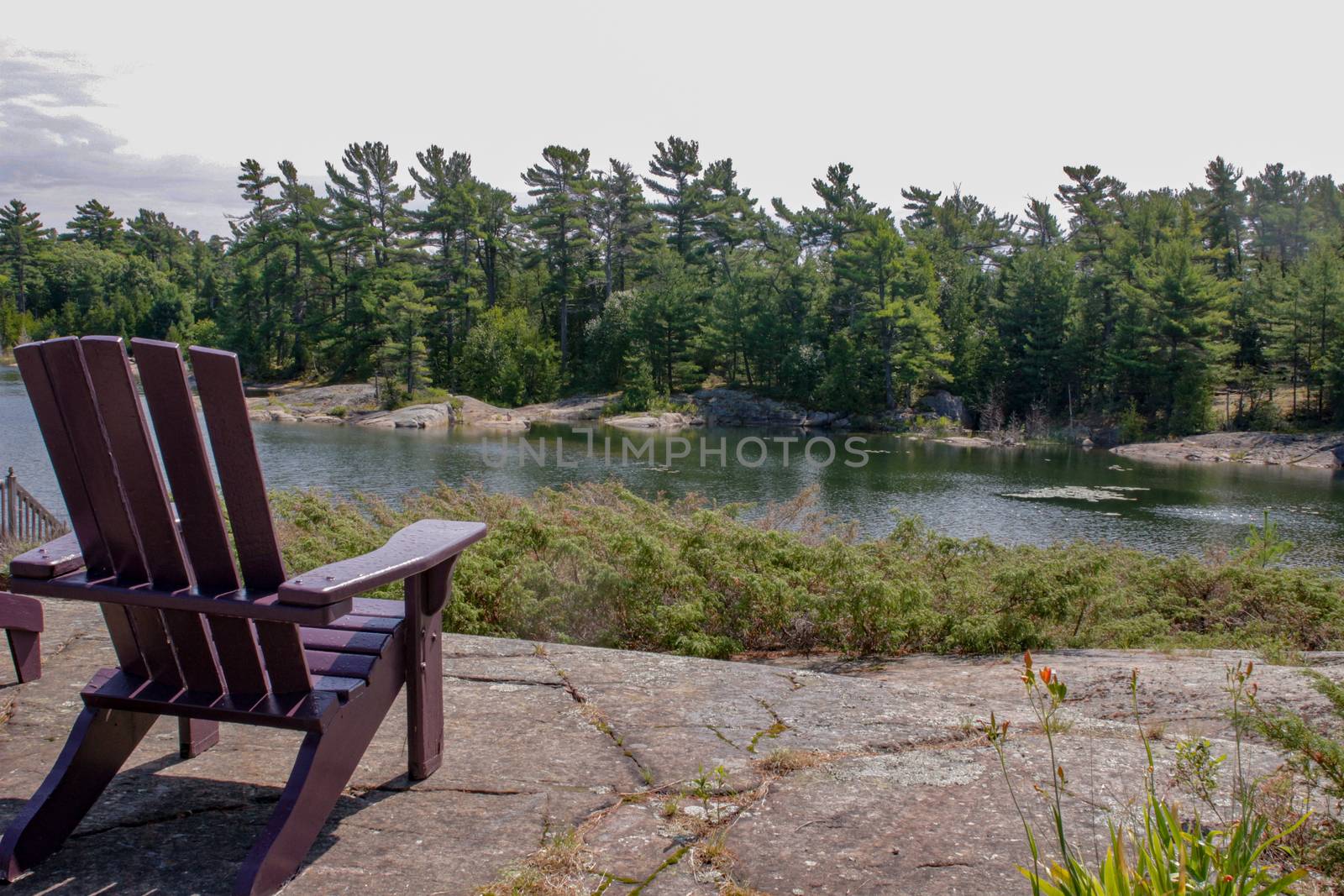 Two black Muskoka chairs sitting on a wood dock facing a lake. Across the calm water is a white cottage nestled among green trees. Canada flag is visible by mynewturtle1