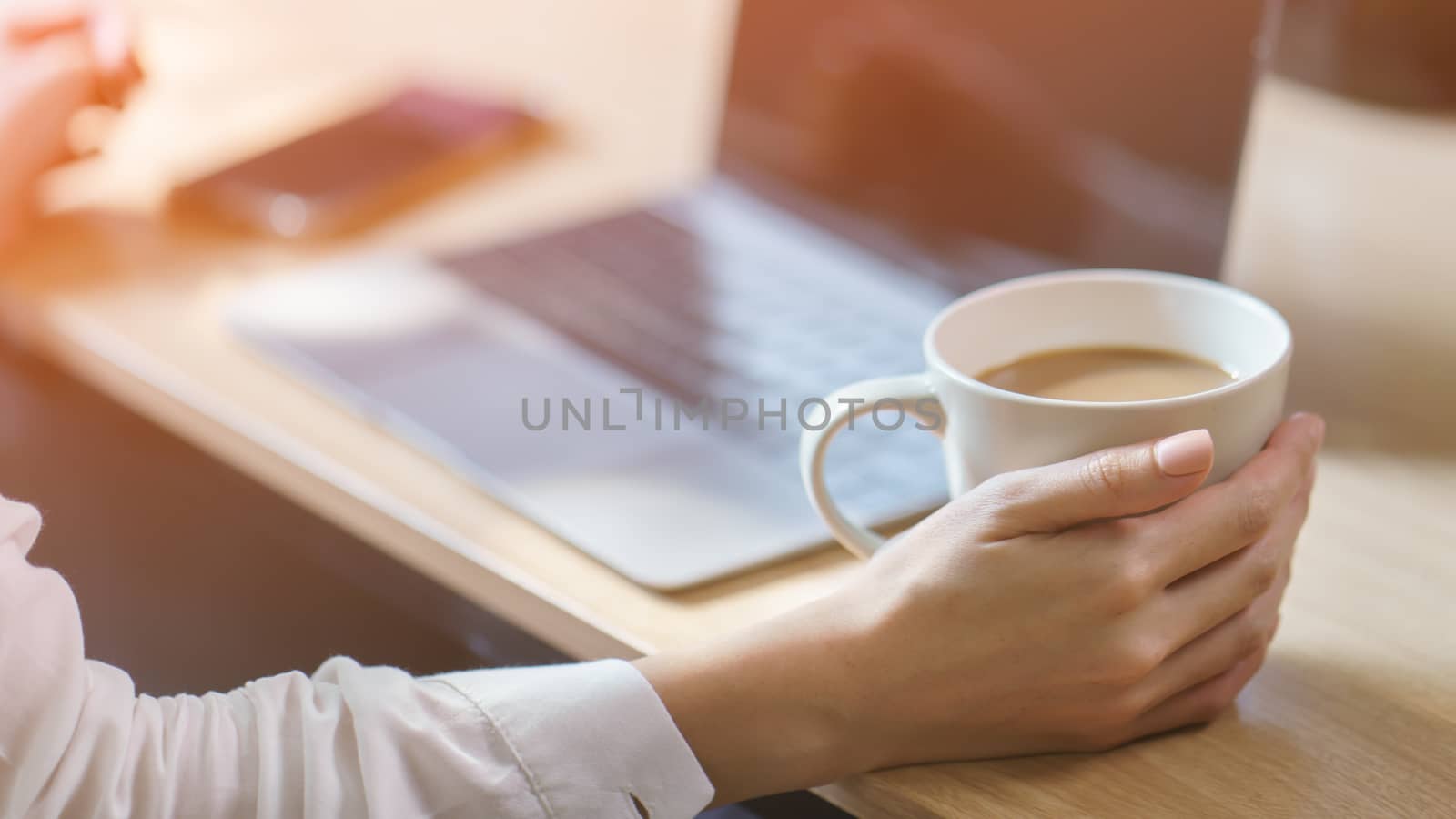 Woman hand on work desk with a laptop computer, a cup of coffee, with morning sunlight.