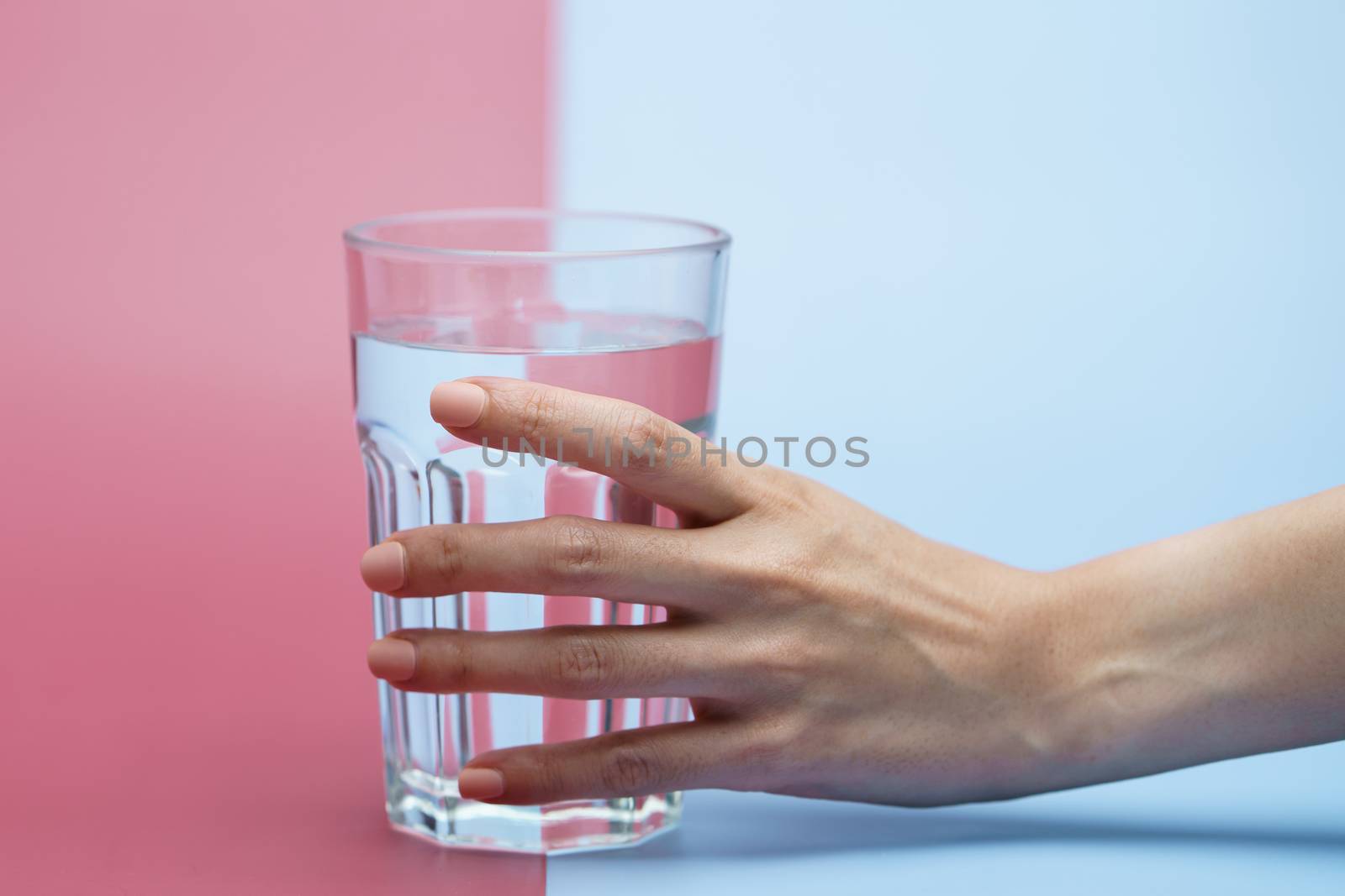 Hand holding transparent glass of water, clean drinking water in a clear glass on two tone (pink and blue) background