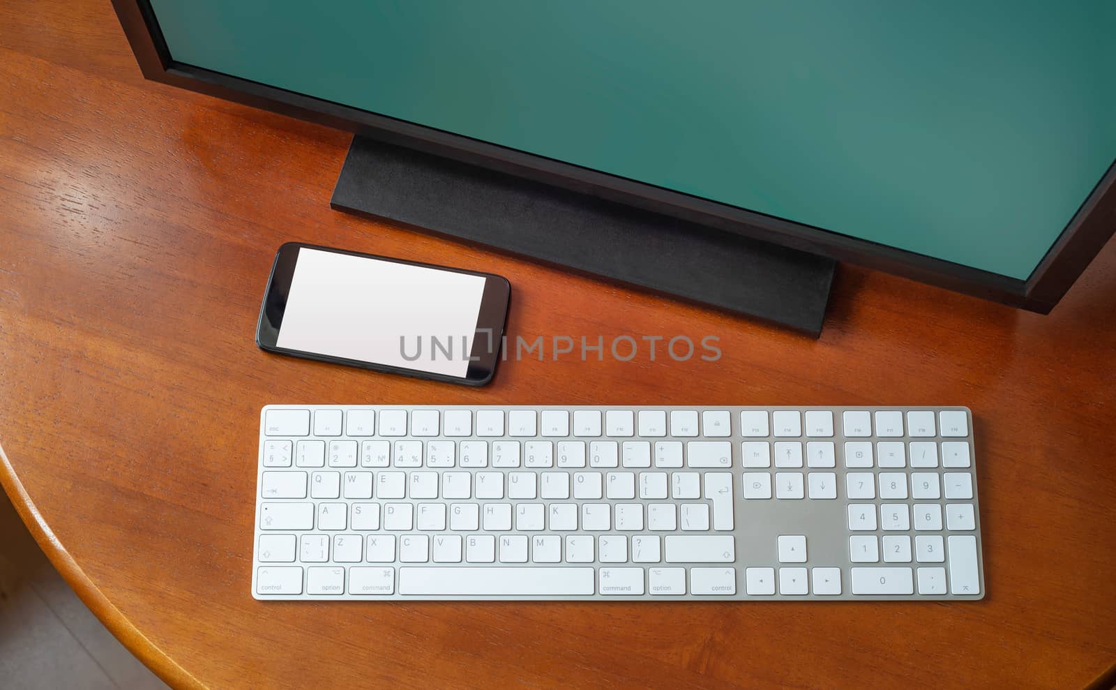 A computer monitor, a digital wireless keyboard and a black smartphone on a round wooden desk