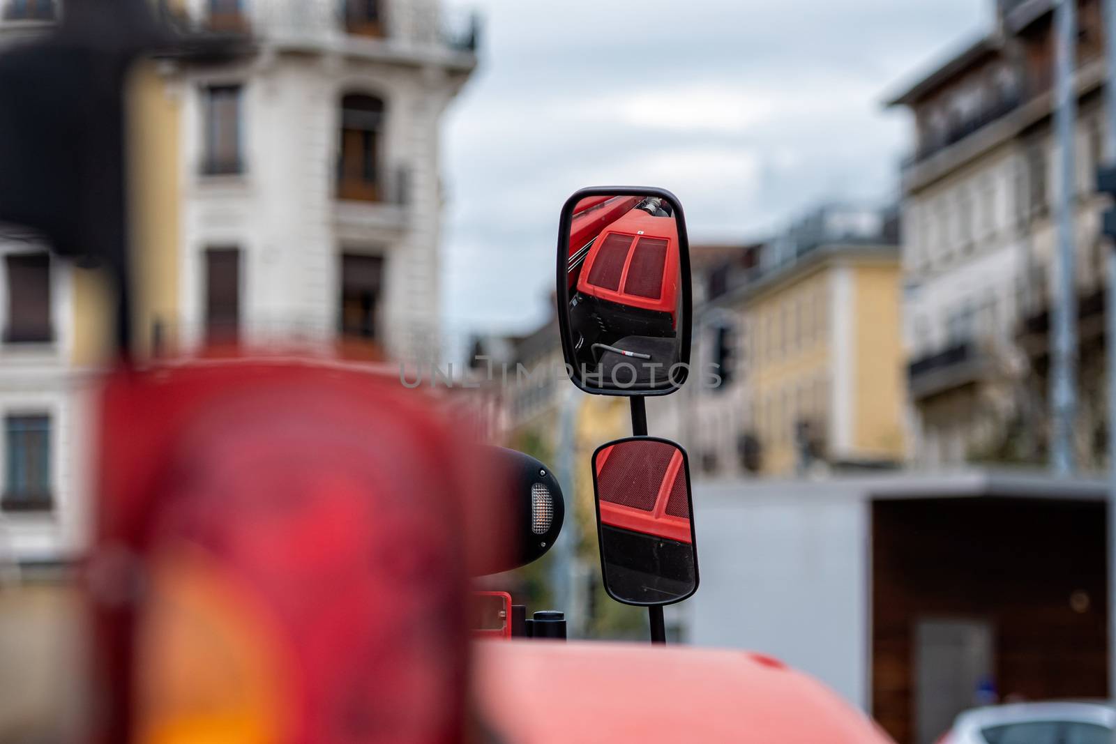 Close-up of tractor side mirror on defocused city background. The mirror shows the red hood of the tractor - image