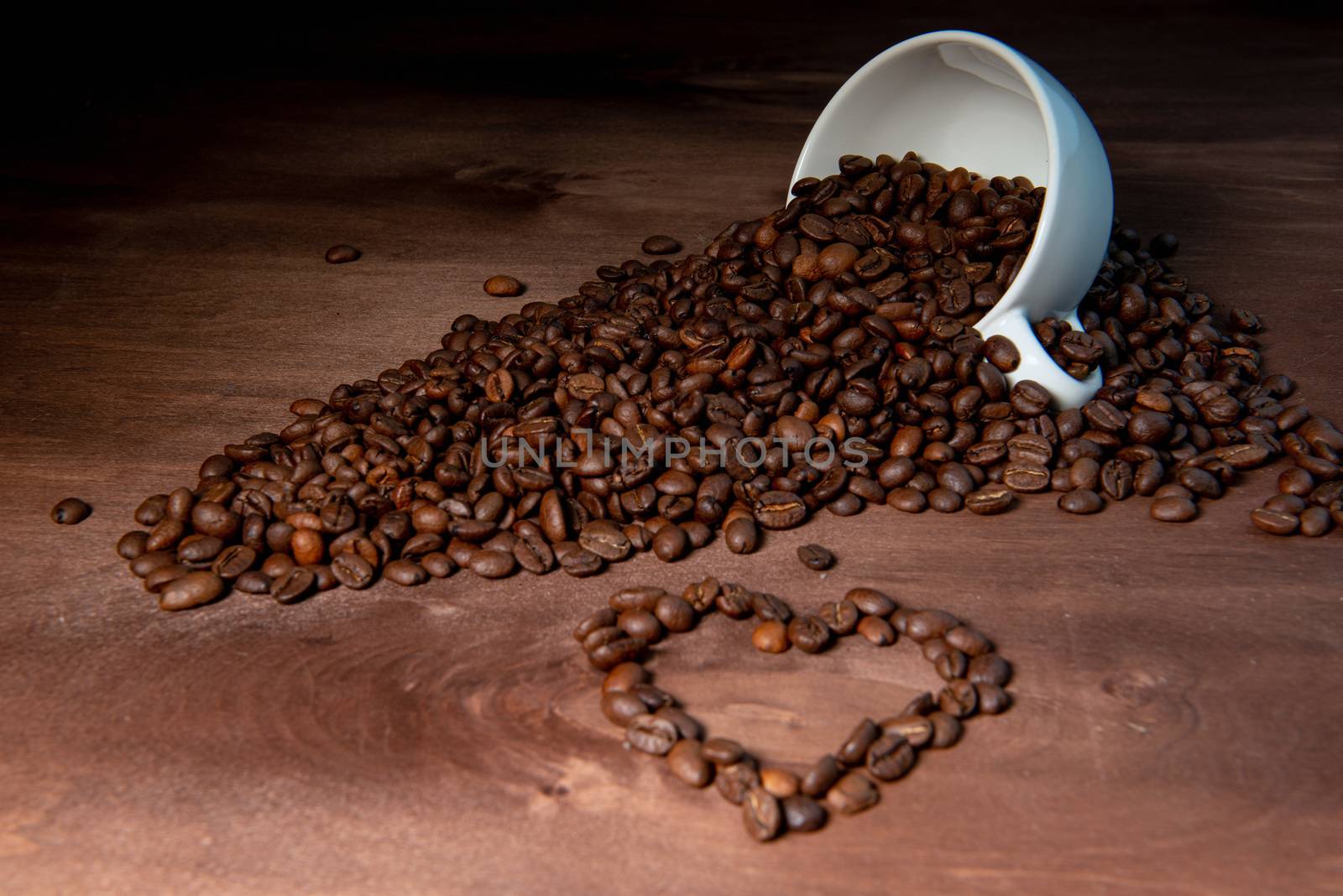 Coffee bean pouring out of a white mug on wooden table, Heart shape from coffee beans in the foreground - image