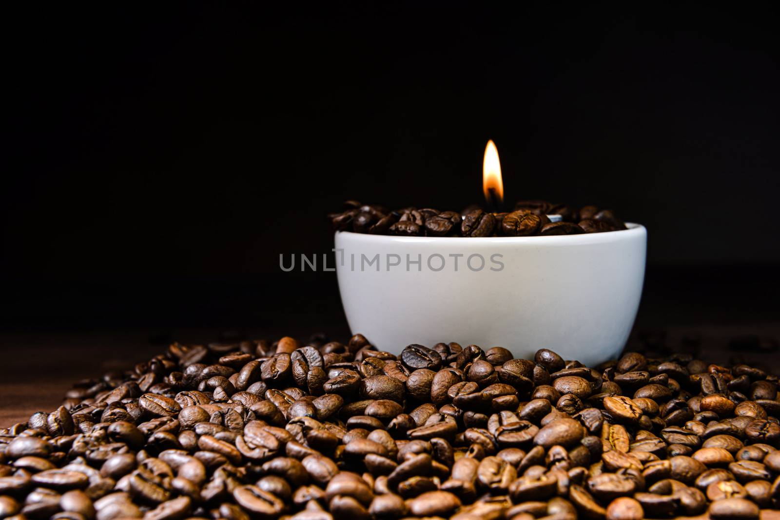 White coffee cup full of coffee beans and burning candle on top of coffee bean stack - image