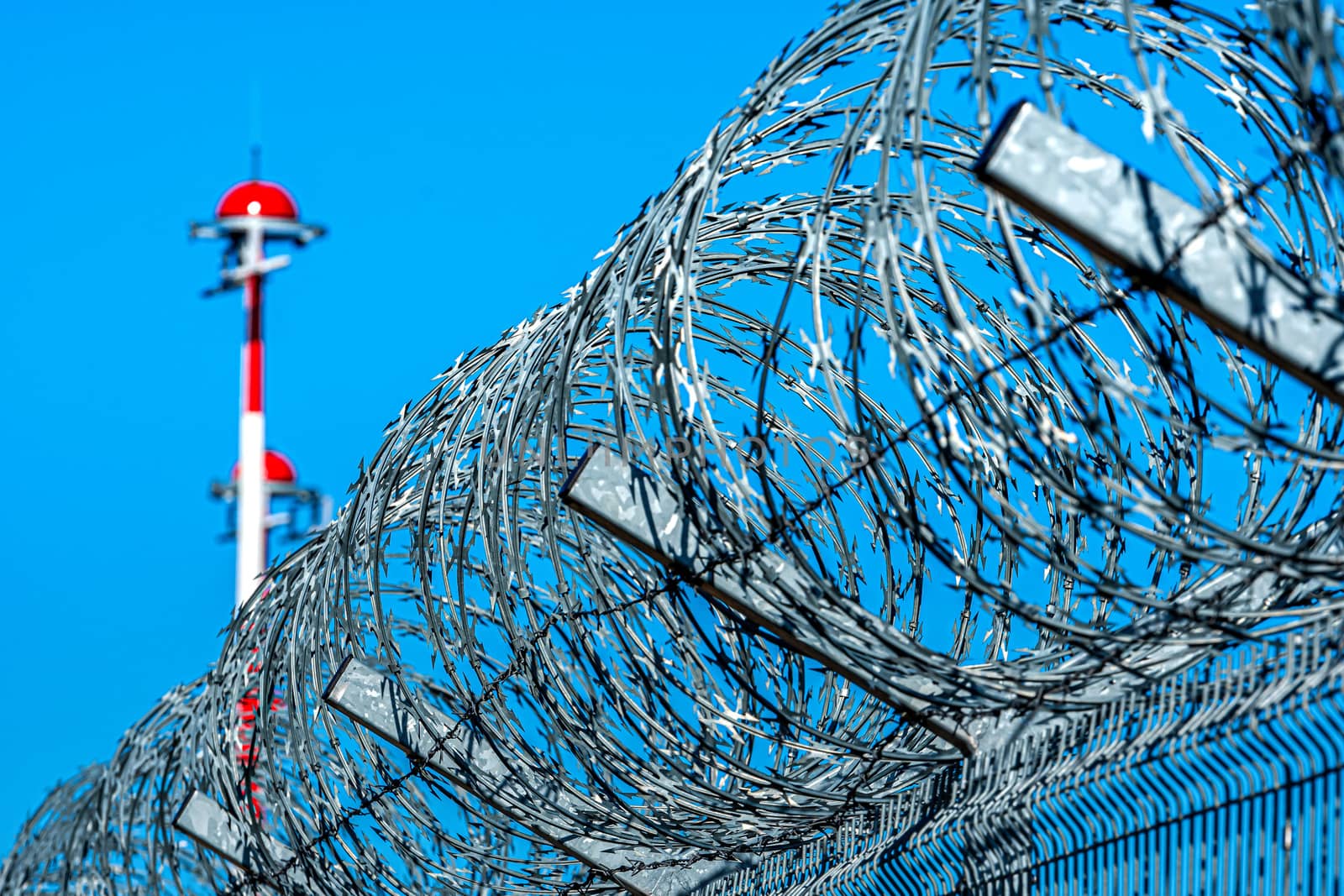 fence with barbed wire and row of the airport lamp posts with alternate red and white painting against blue sky, security concept