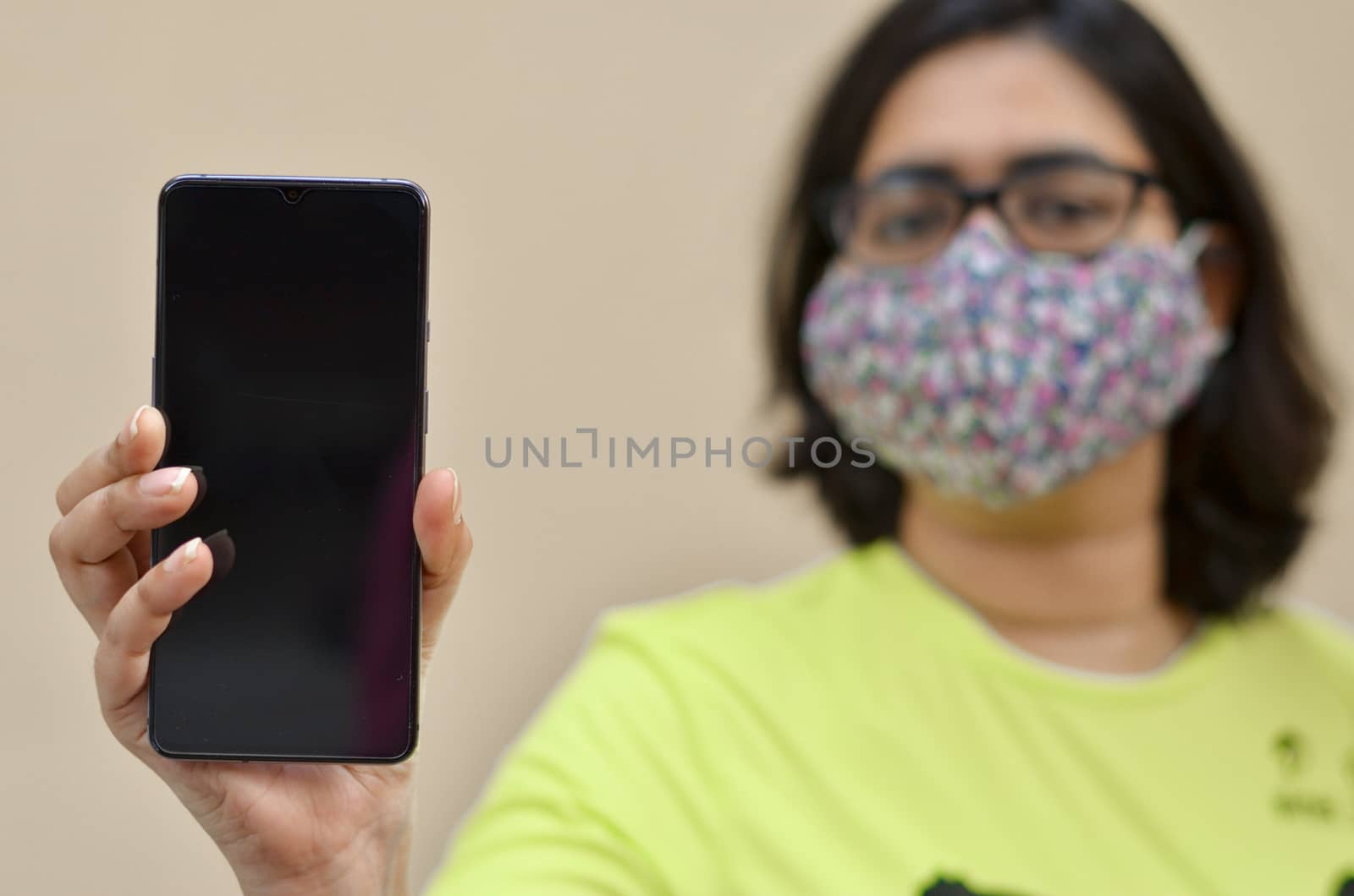 Young woman wearing surgical face mask showing her blank mobile phone black screen against yellow background which can be used for copy space. Concept Corona virus (Covid-19) disease pandemic by jayantbahel