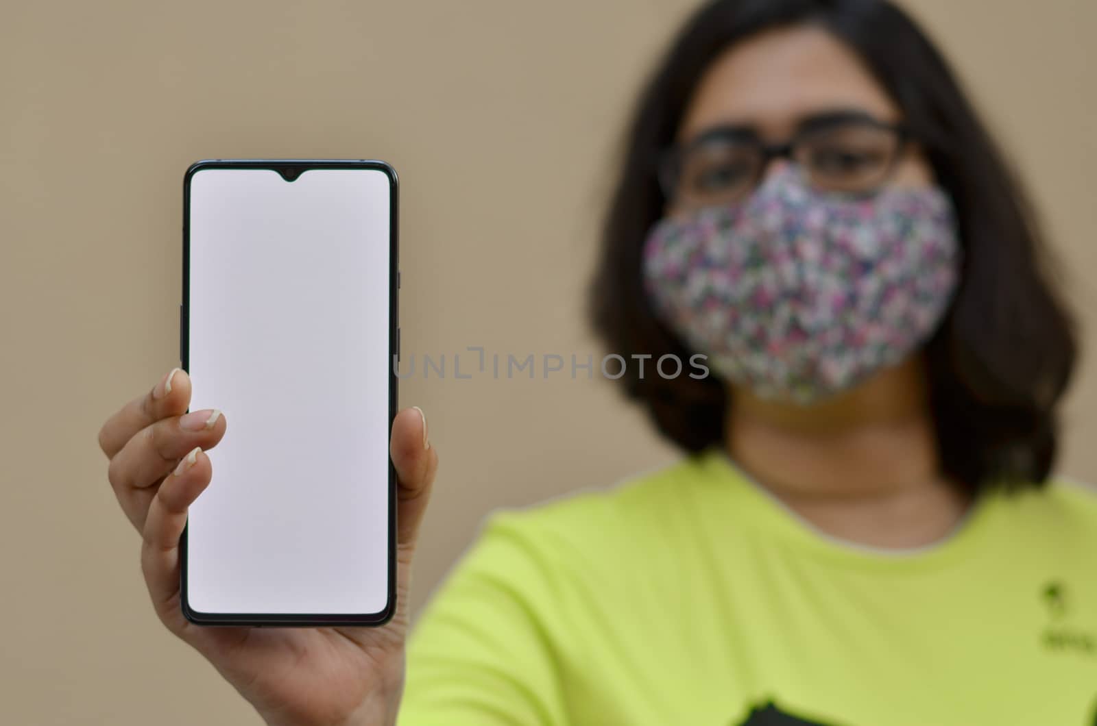 Young woman wearing surgical face mask showing her blank mobile phone white screen against yellow background which can be used for copy space. Concept Corona virus (Covid-19) disease pandemic by jayantbahel