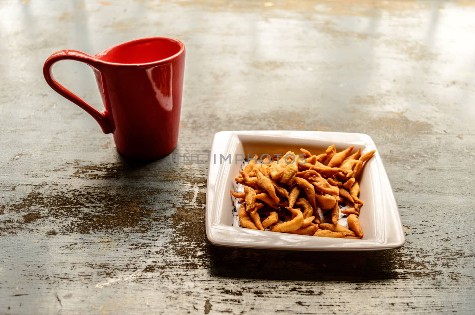 Close up Red Color Coffee mug and white plate full of Namkeen Bhujia snack cookies on old rustic floor. Food and drink background. Morning sunlight coming from window.