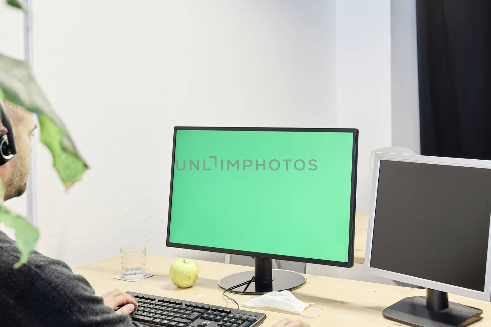 Man at his workplace with a computer and a green screen, mask, apple and glass of water on his desktop