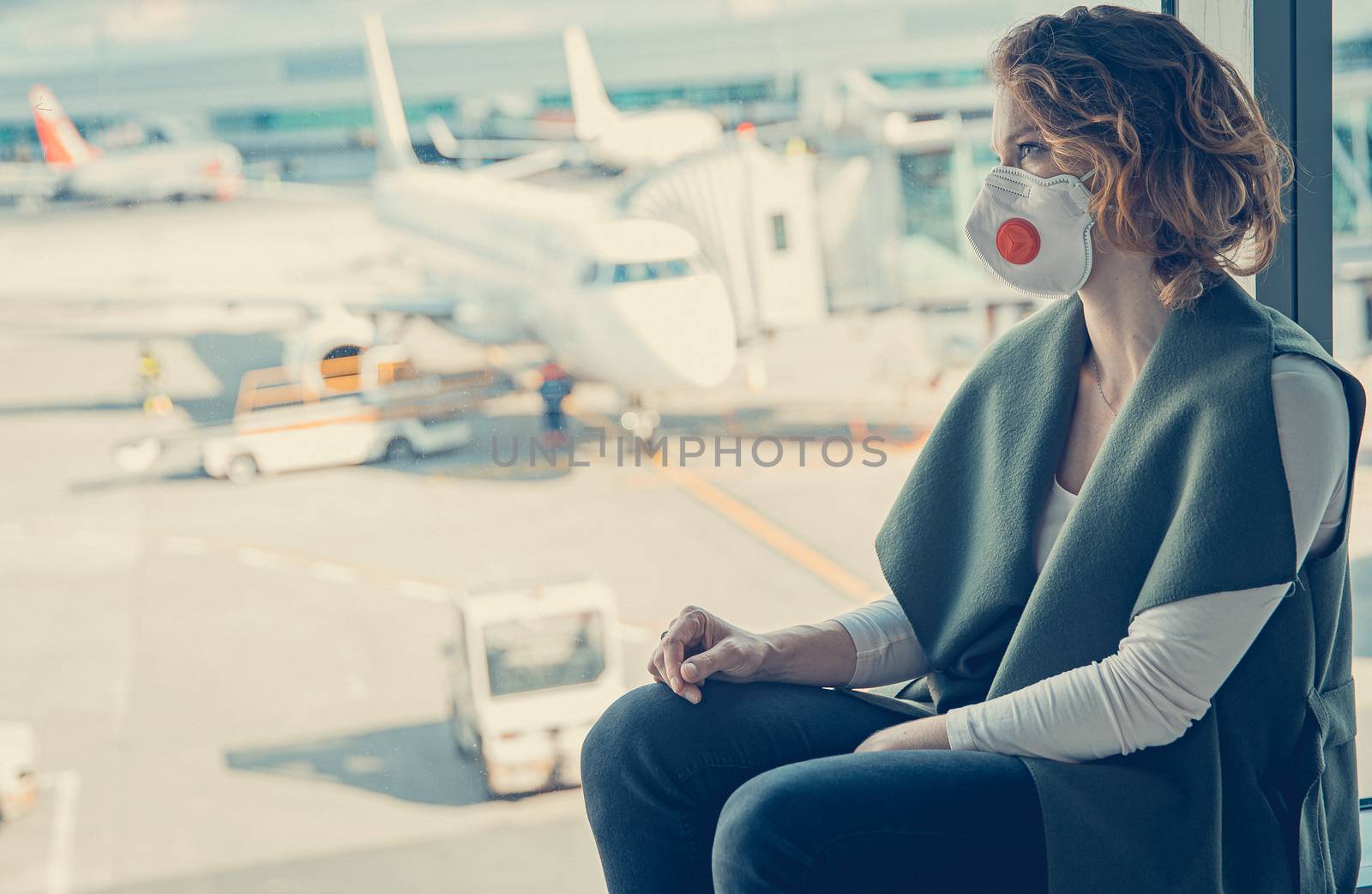 Woman with a mask on her mouth protects against the virus. She looks sad through the window at the airport on planes. Aerial connections canceled due to a coronavirus.