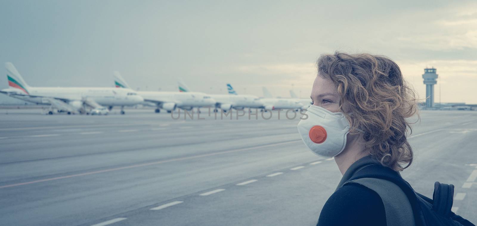 Woman with a mask on her mouth protects against the virus. She looks sad through the window at the airport on planes. Aerial connections canceled due to a coronavirus.