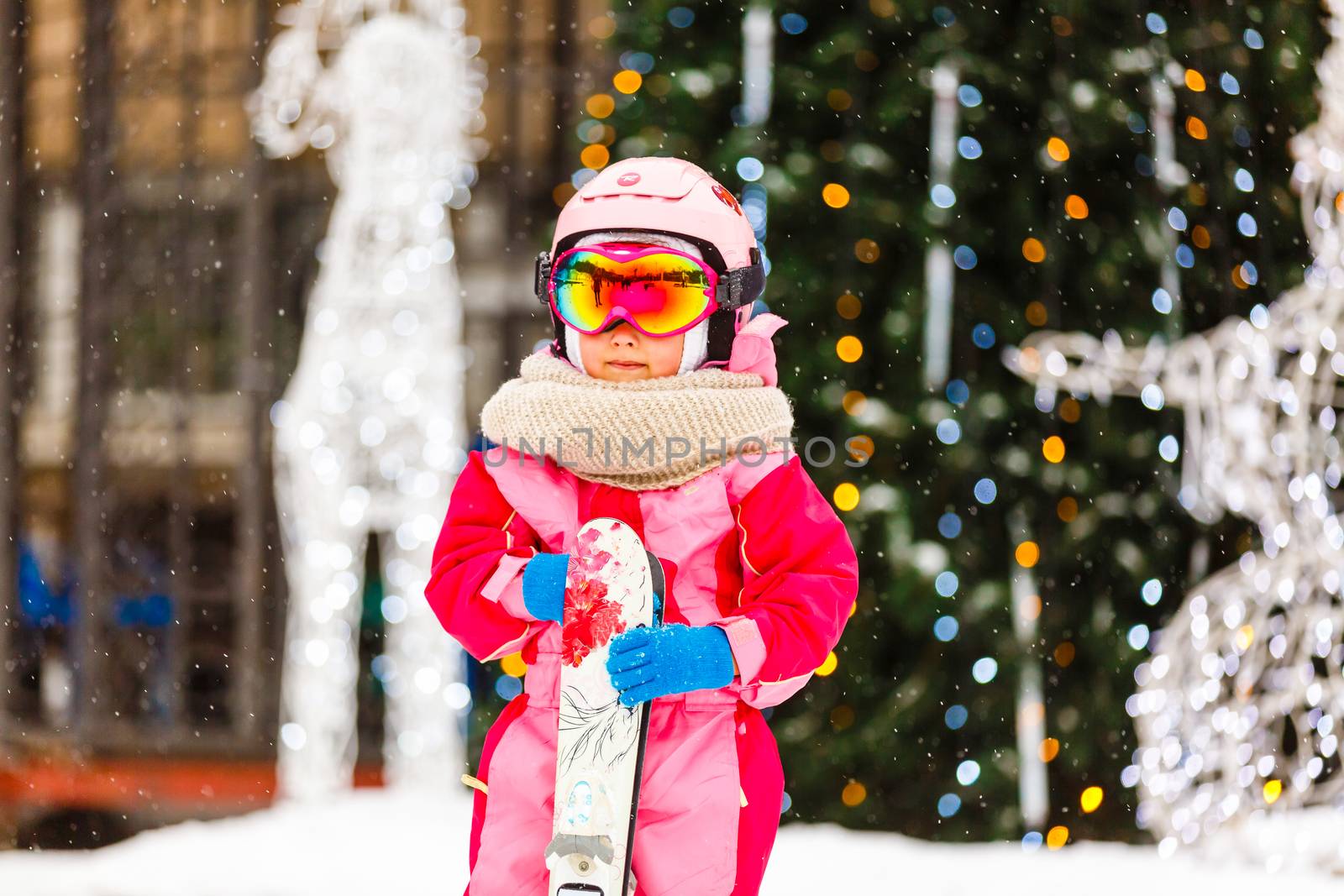 Little girl wearing goggles and helmet on a winter vacation outdoors holding skis behind looking camera smiling excited close-up blurred background