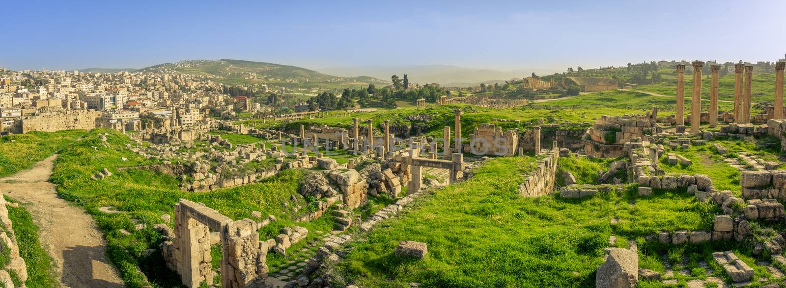 Panorama overview of the Roman site of Gerasa, Jerash, Jordan, with ruins, pillars and remains of the old city clearly visible. Green grass and blue sky on a sunny day in spring.