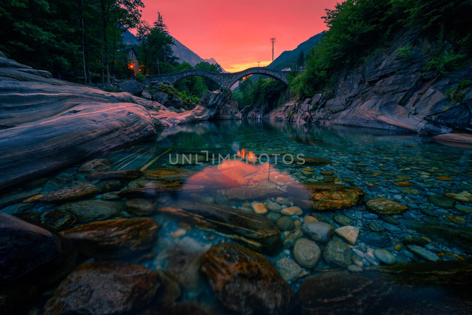 Colorful sunset above the double arch stone bridge at Ponte dei Salti in Lavertezzo, Canton Tessin, Switzerland, with the clear water of Verzasca river in the foreground.