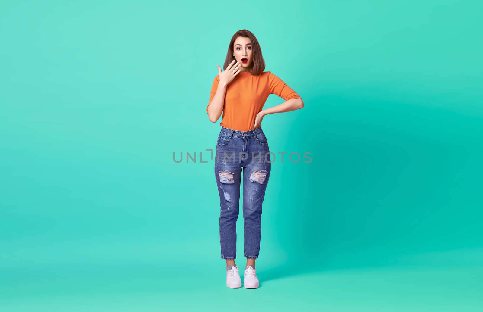 Shocked excited beautiful woman with mouth open wearing casual orange t-shirt over blue background.