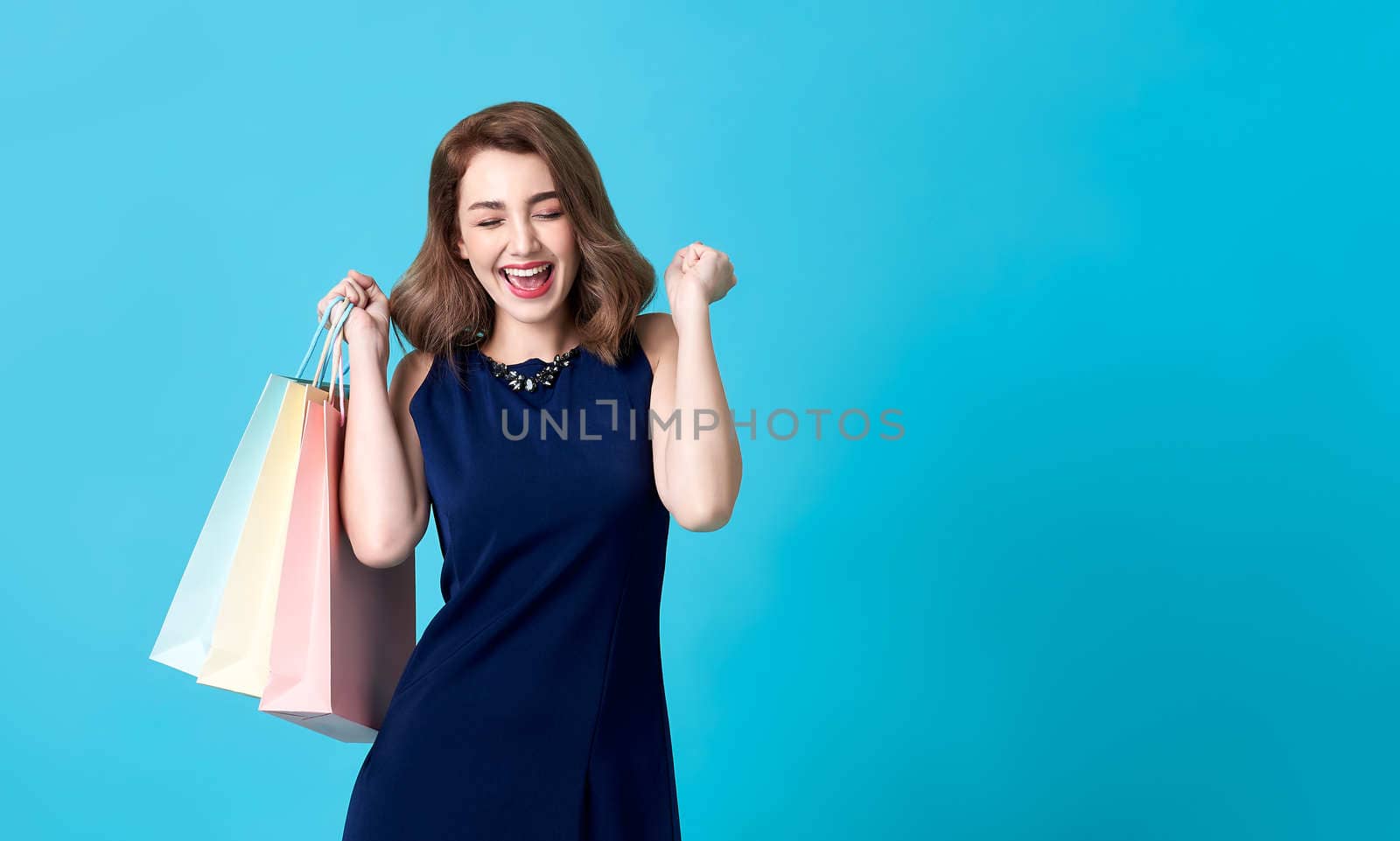 Portrait of an excited beautiful woman wearing blue dress and holding shopping bags isolated on blue background.