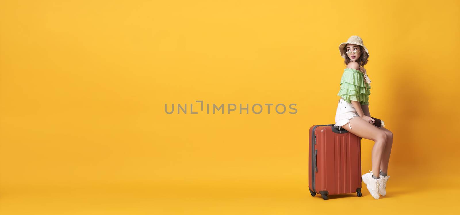 Cheerful young woman dressed in summer clothes holding passport with suitcase over yellow banner background with copy space.