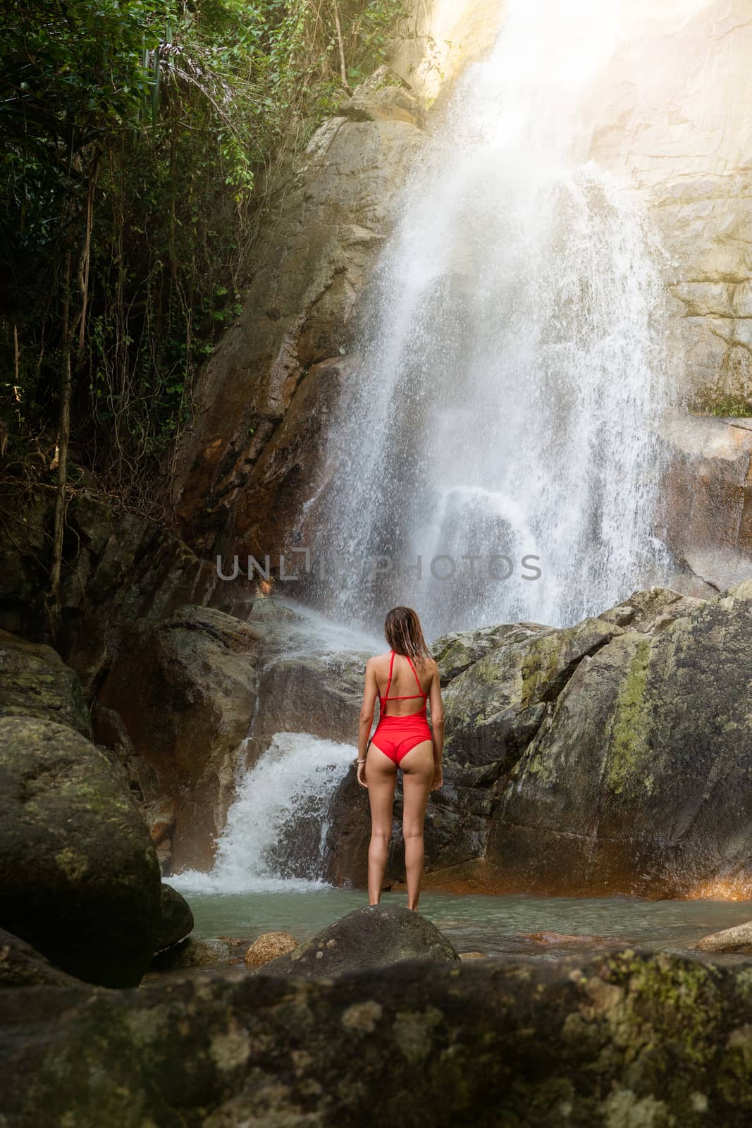 Beautiful young slim blonde woman with curly hair, wearing in red swimsuit is enjoy in lagoon of huge tropical waterfall in jungle. Travel concept. Sexy woman wearing red swimsuit in the jungle