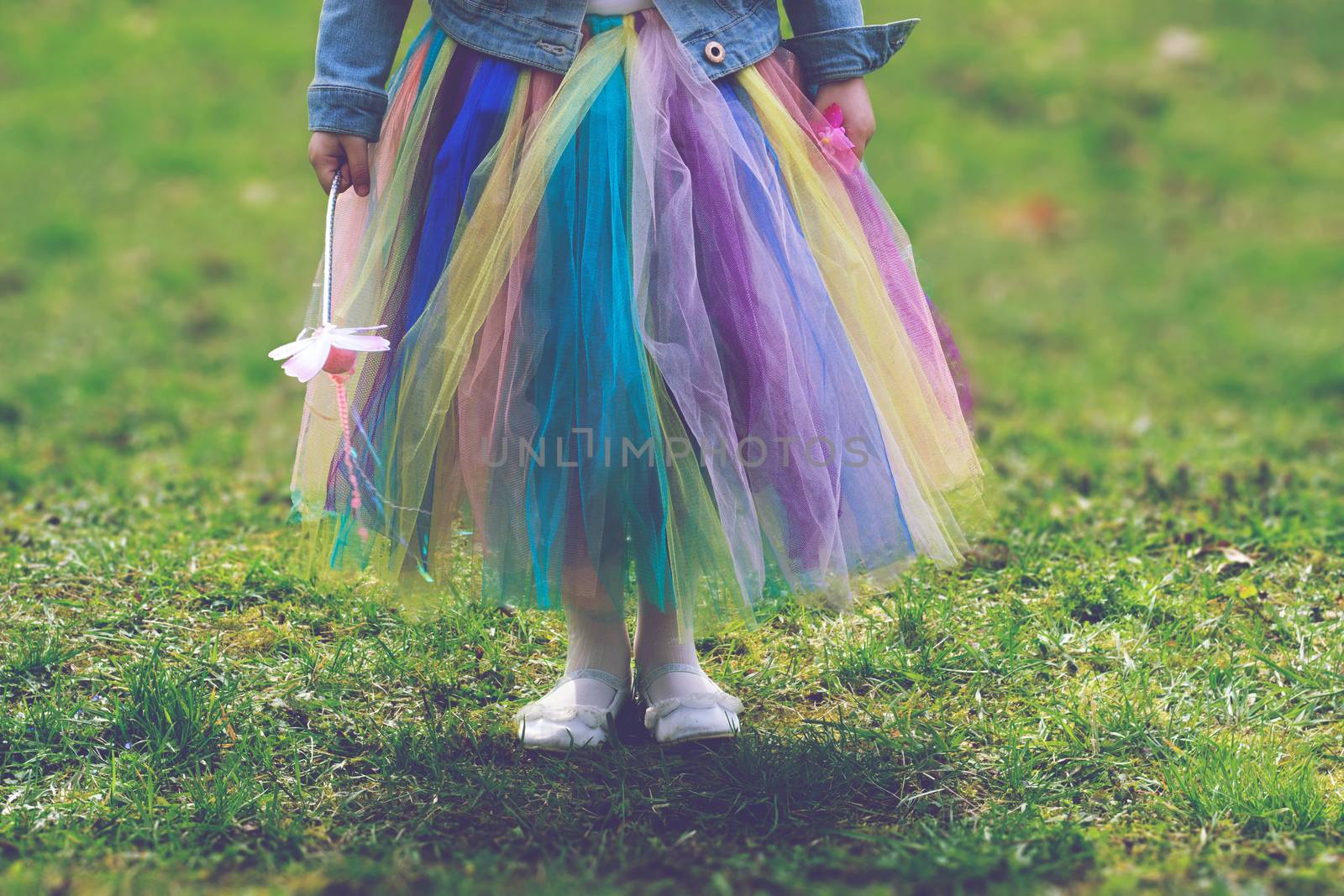Little girl in colorful tutu skirt standing on green grass while holding magic wand