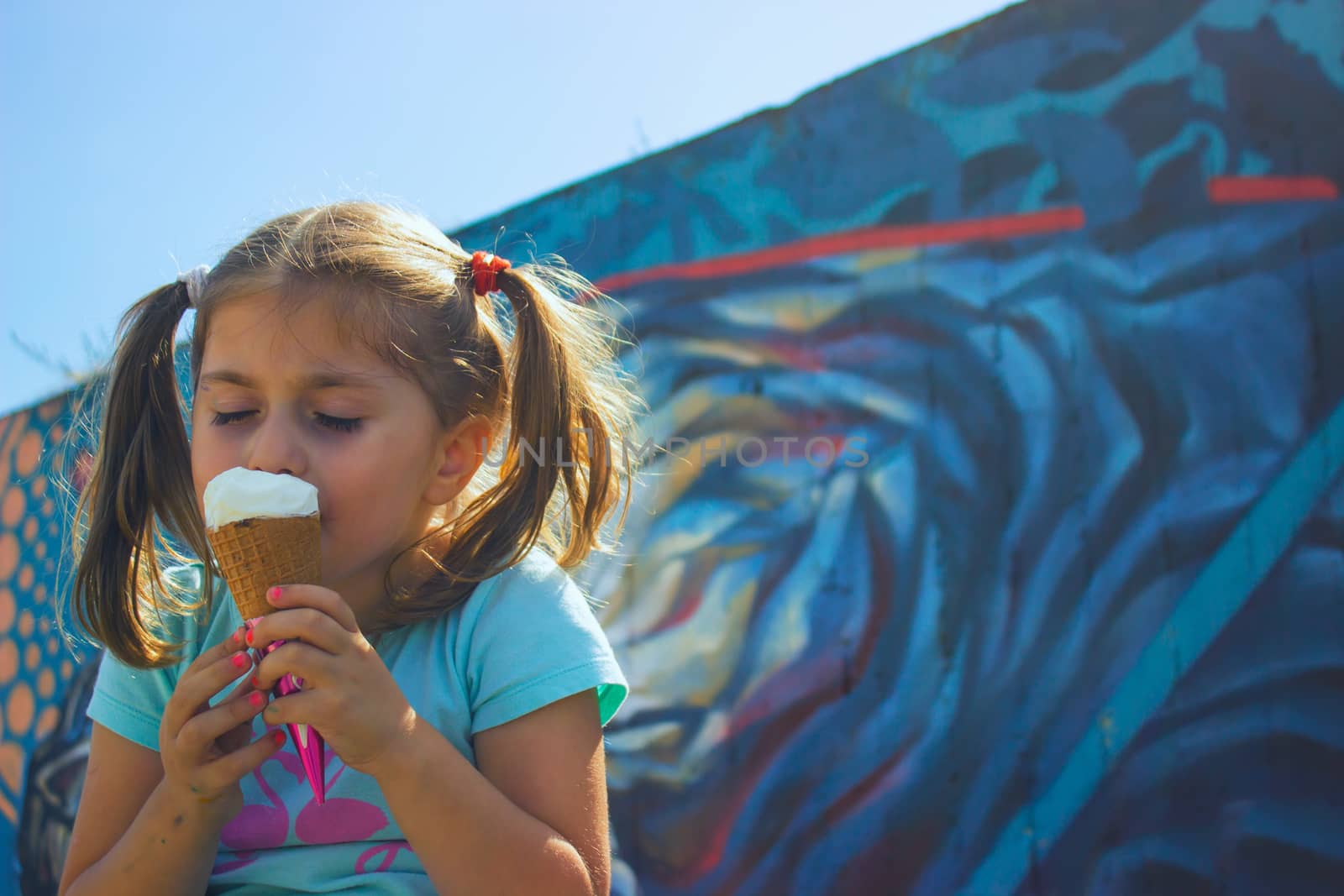 Little girl eating ice cream with her eyes closed, against blue wall