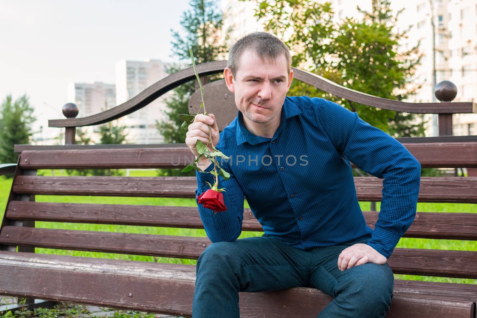 A man with a rose in his hands sits on a bench in a park with a displeased face
