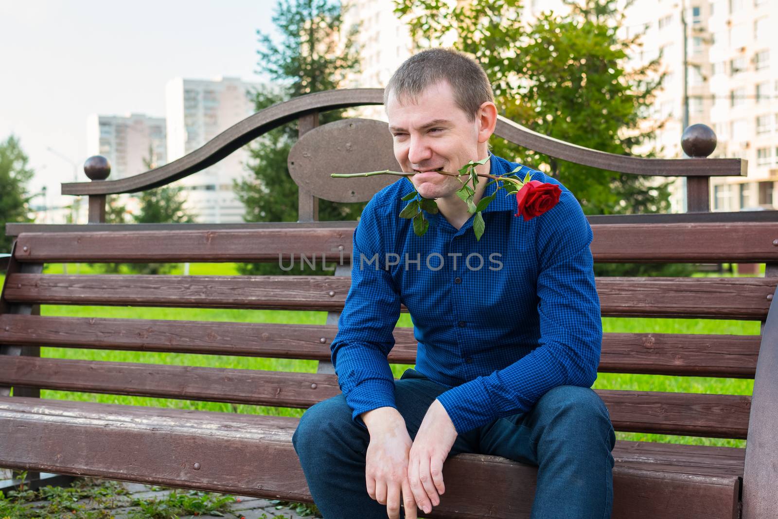 A man with a rose in his teeth sits on a park bench and is looking for single ladies