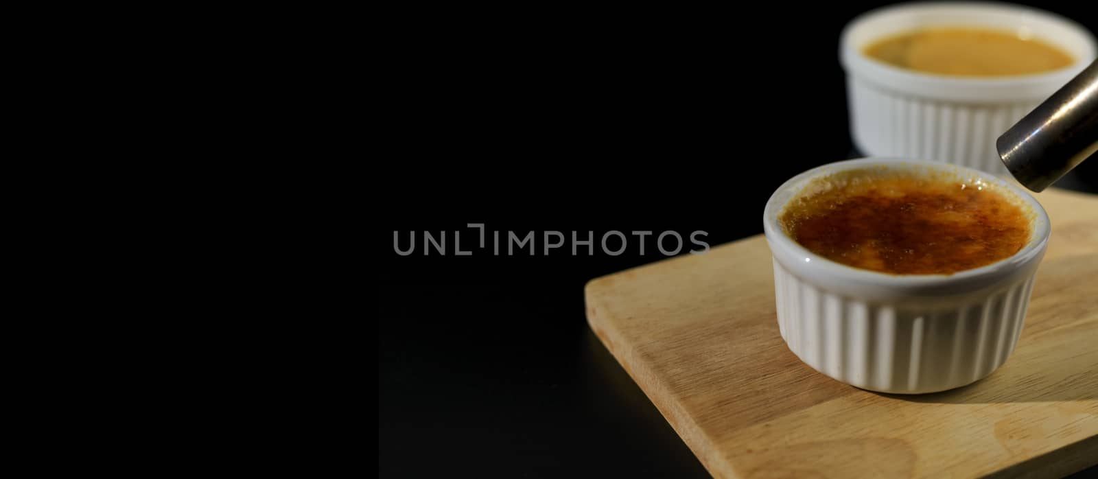 Close up homemade creme brulee in white ramekin  with butane torch burn sugar on top  on black background with copy space. French dessert creme brulee