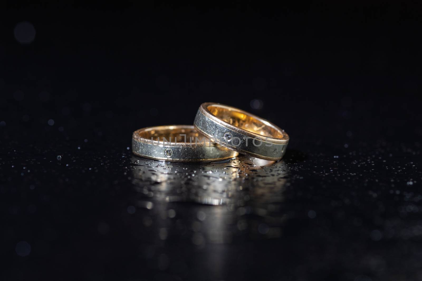 Wedding rings lying on dark surface shining with light close up macro. Water splashes