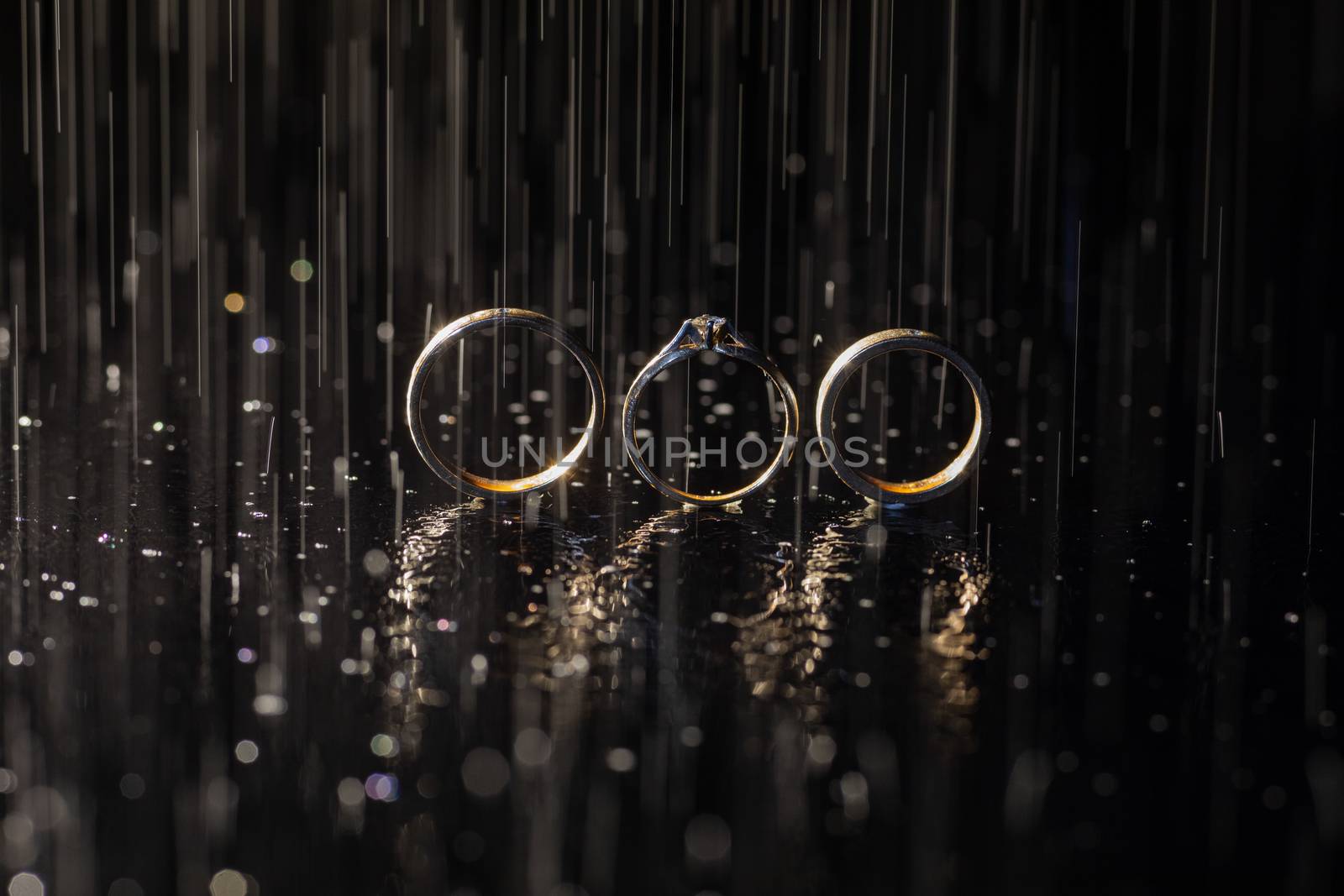 Wedding and engagement rings lying on dark surface shining with light close up macro. Water splashes. Rain