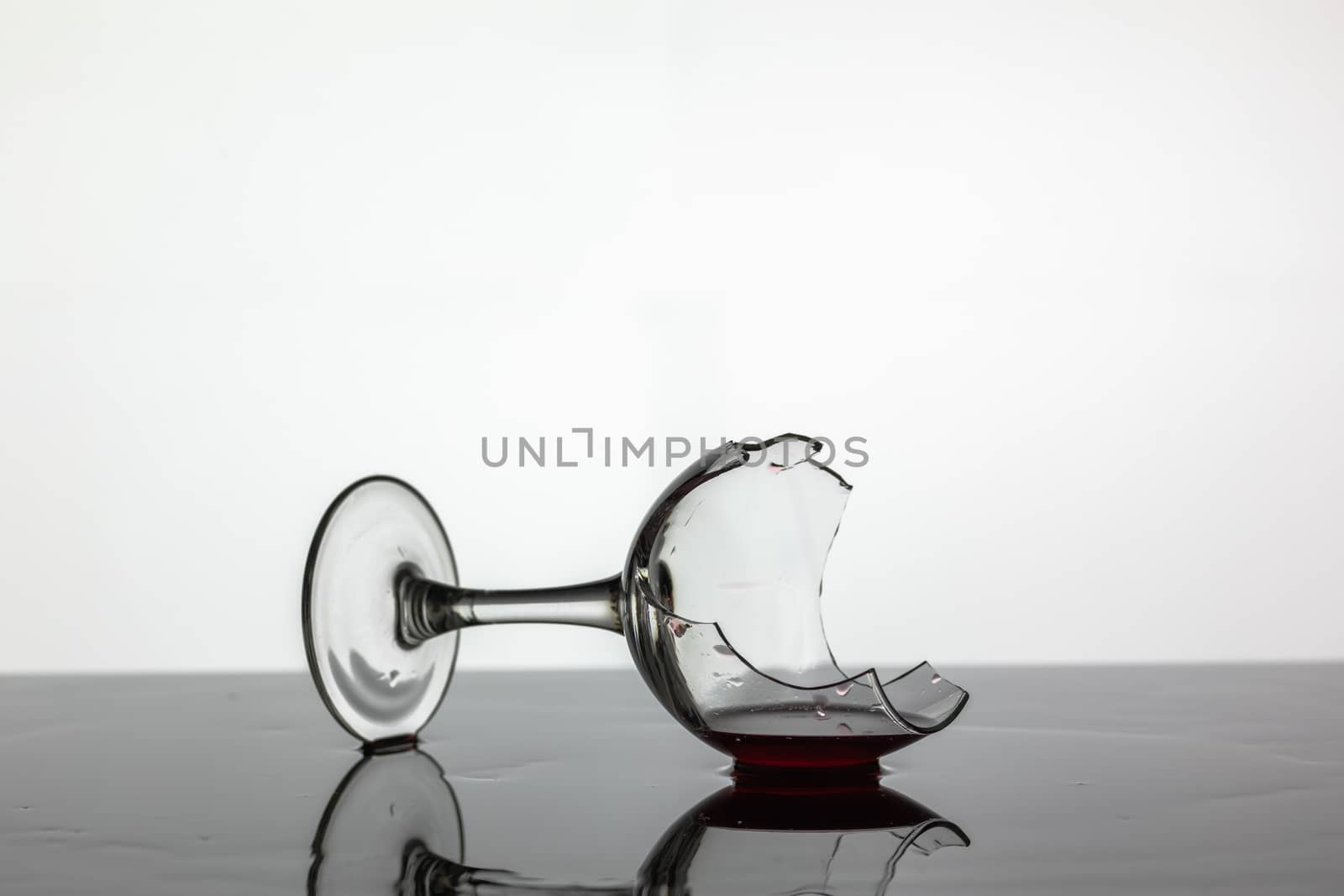 Broken wine glass with red wine which is laying on the wet surface. White background