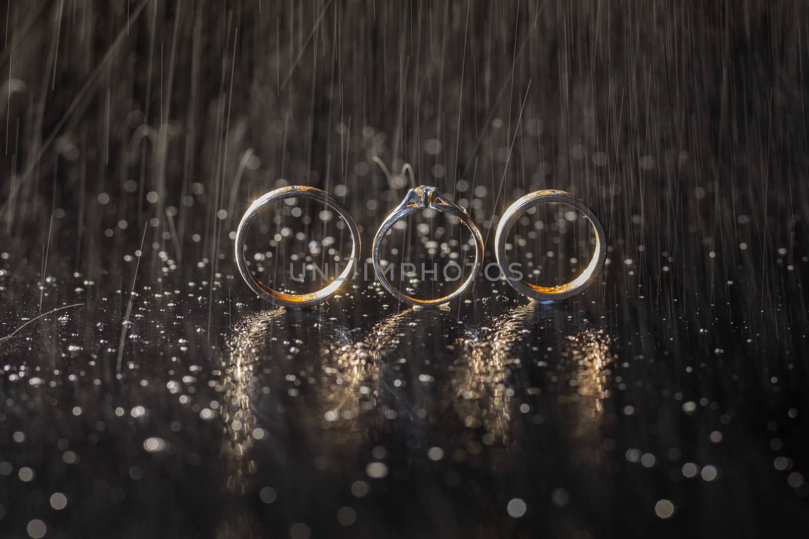 Wedding and engagement rings lying on dark surface shining with light close up macro. Water splashes. Rain