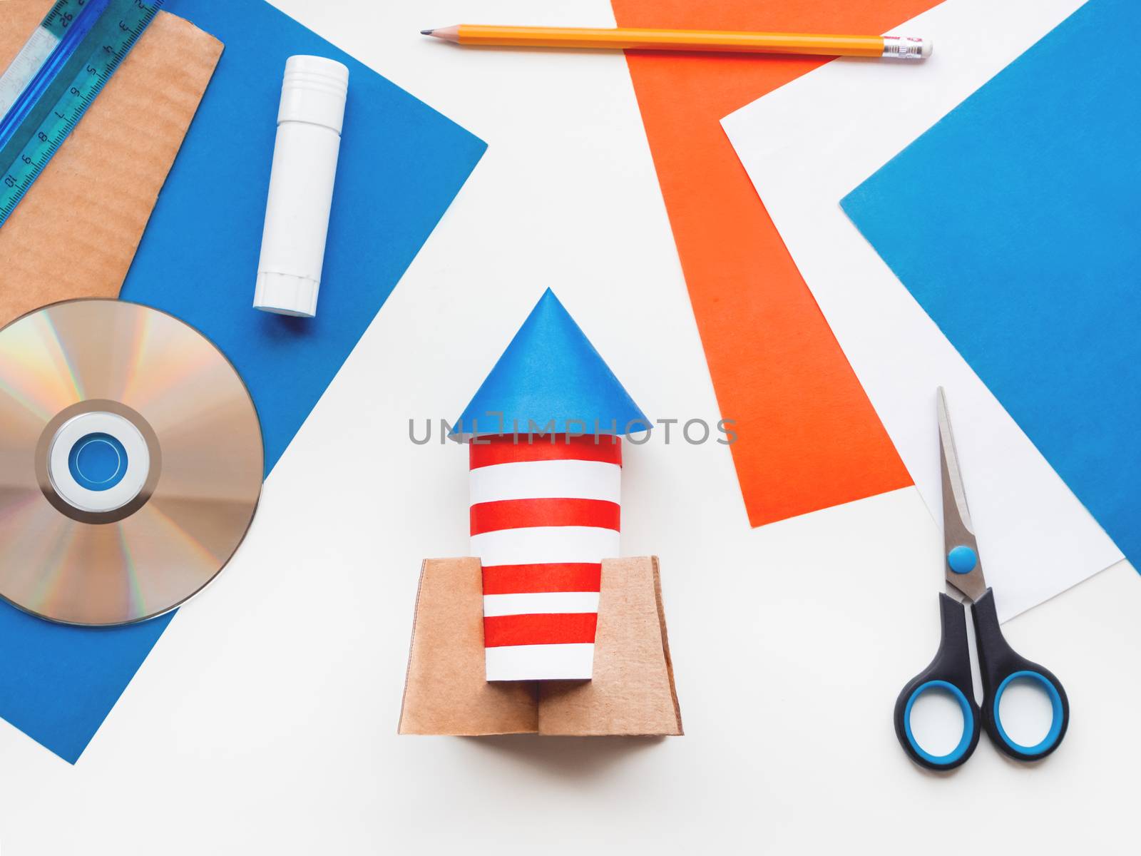 Child made colorful rocket out of colored paper. Also he needs carton, scissors, glue, CD-disk, pencil and ruler. Red, white and blue handmade spaceship. Making toys from improvised materials.