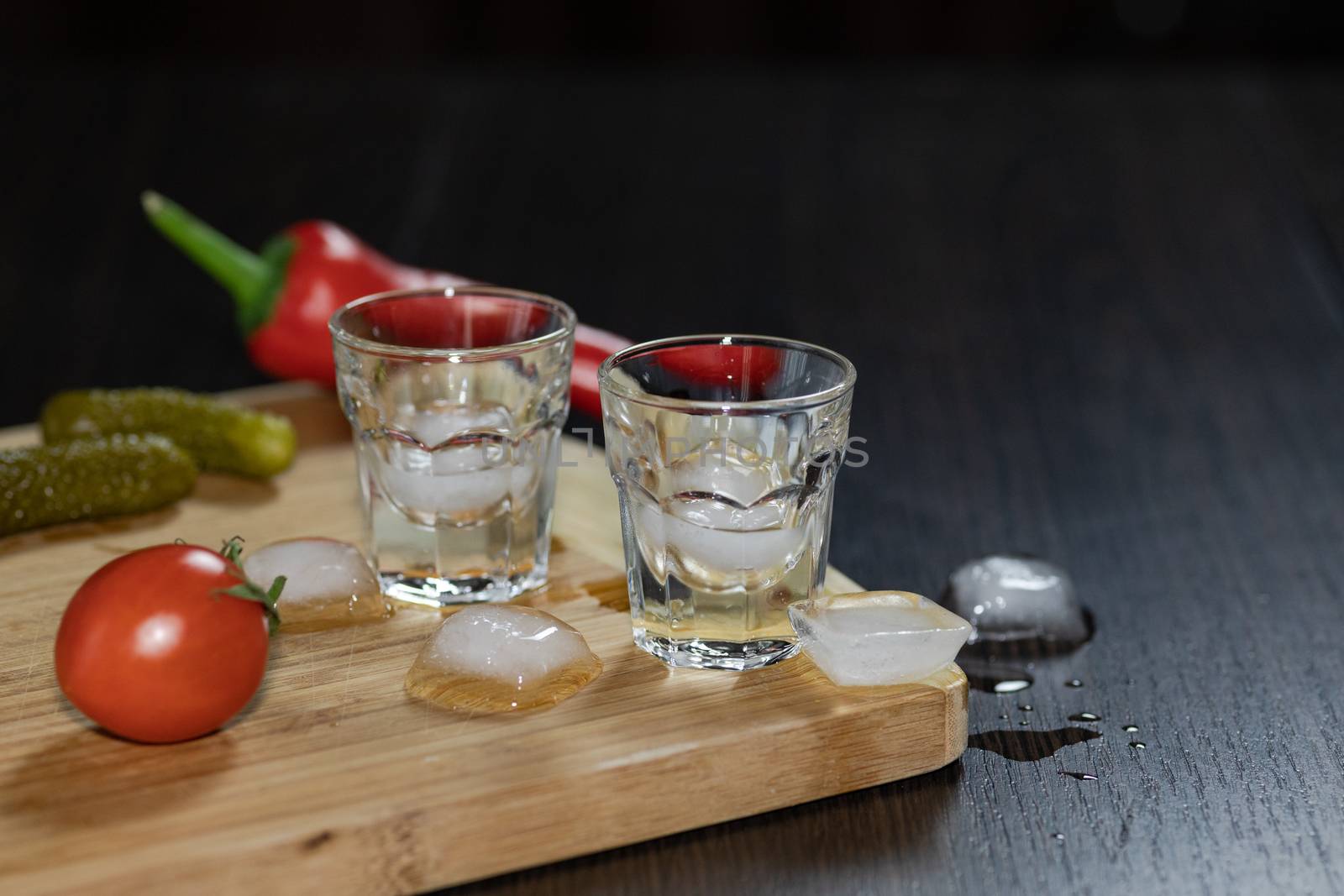 Two shot glasses of vodka on the cutting board with cherry tomatoes, marinated cucumbers, pepper and ice cubes. Black background