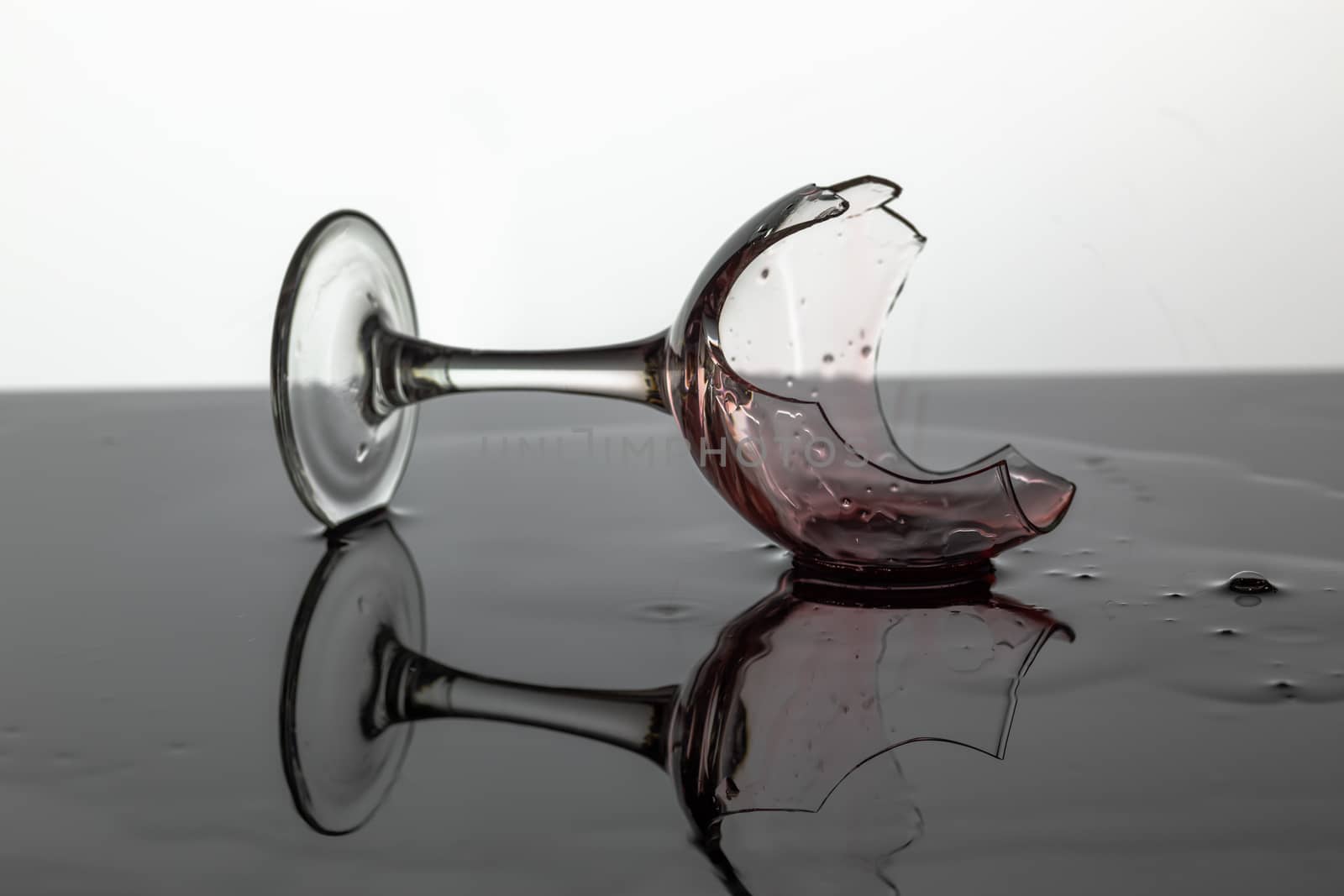 Broken wine glass with red wine which is laying on the wet surface. White background
