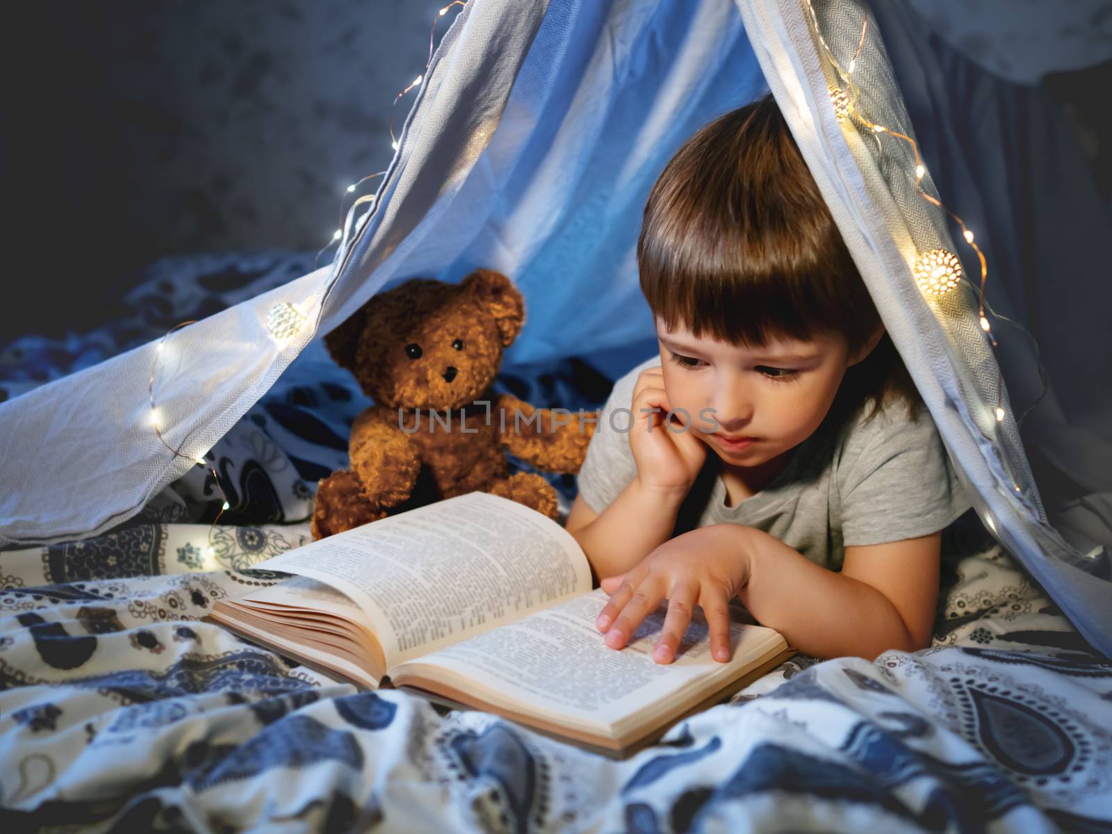 Little boy reads book. Toddler plays in tent made of linen sheet with light bulbs on bed. Cozy evening with favorite book.