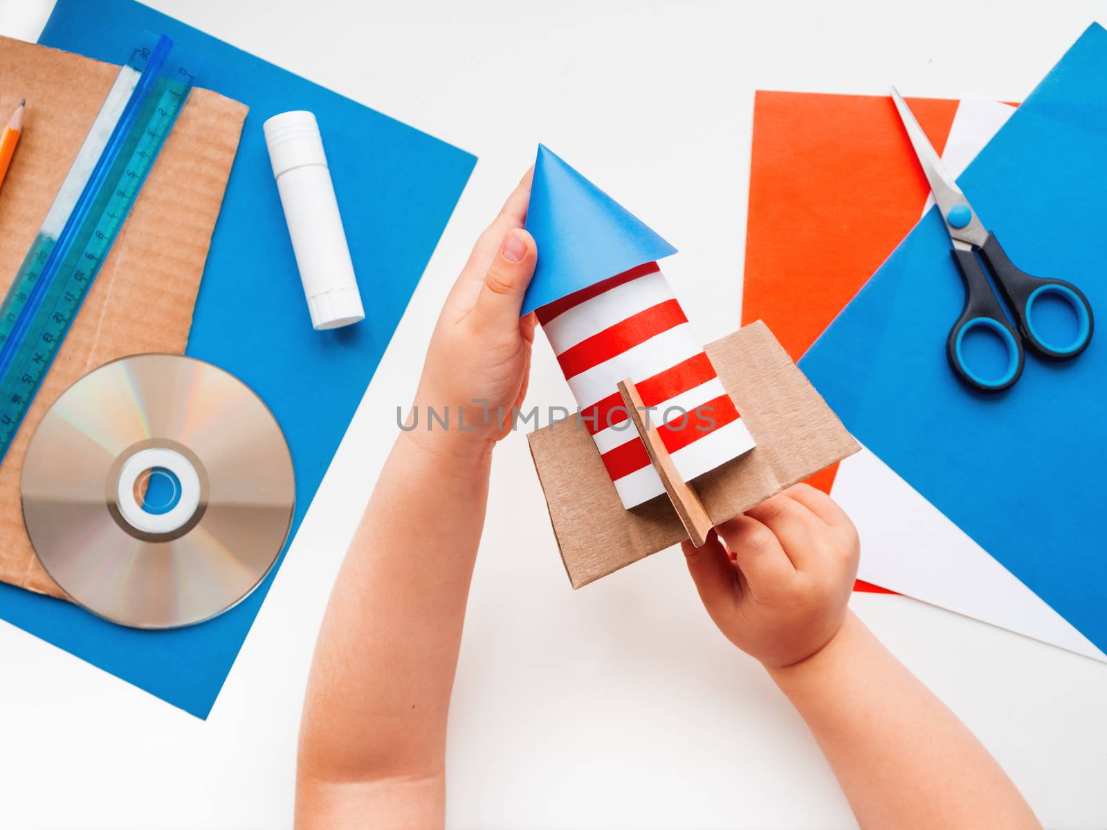 Child makes colorful rocket out of colored paper. Also he needs carton, scissors, glue, CD-disk, pencil and ruler. Red, white and blue handmade spaceship. Making toys from improvised materials.