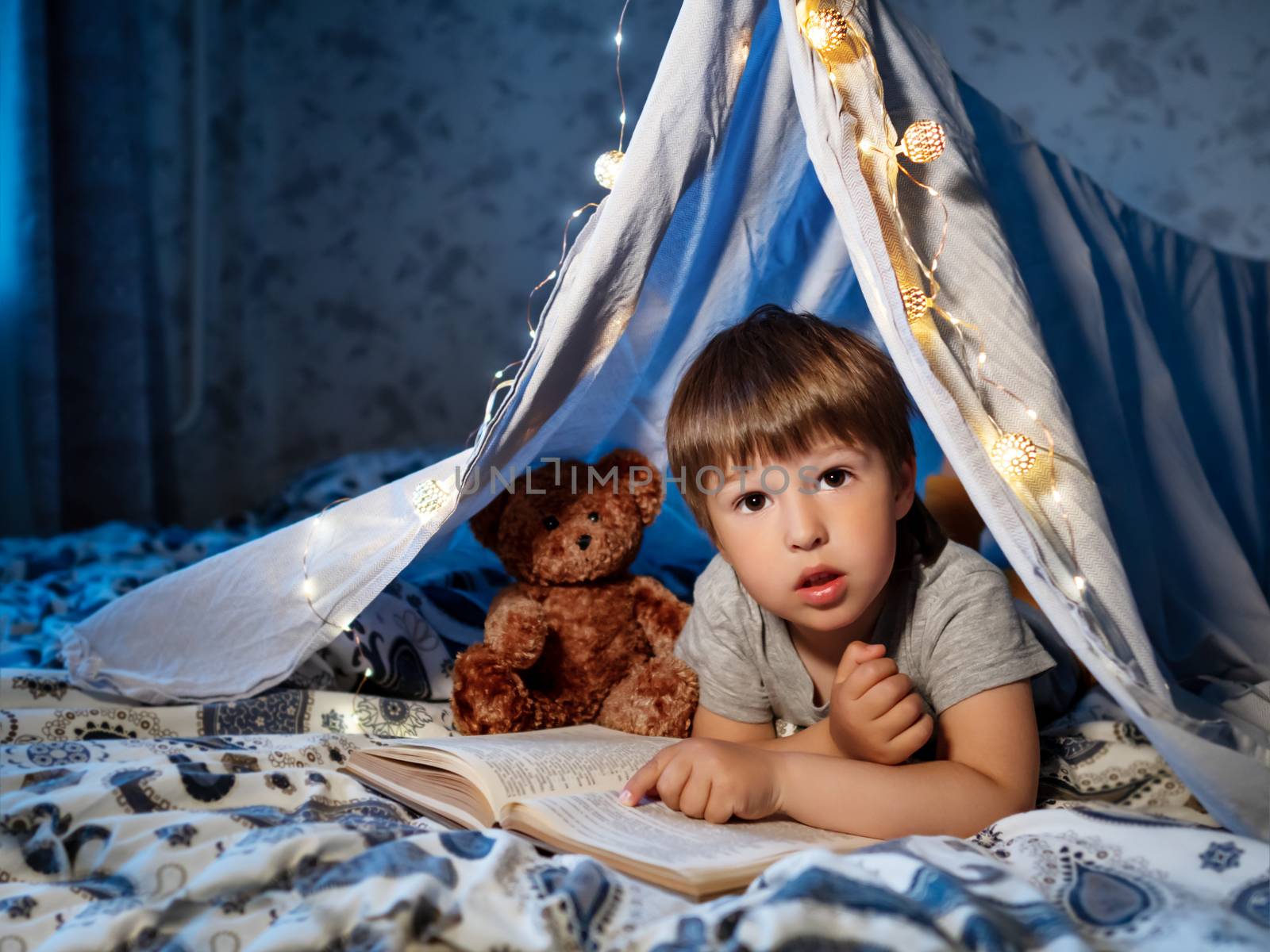 Little boy reads book. Toddler plays in tent made of linen sheet on bed. Cozy evening with favorite book.