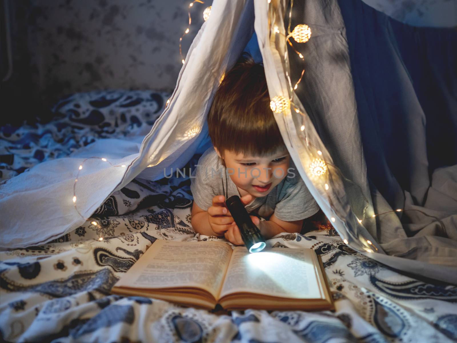 Little boy reads book with pocket light. Toddler plays in tent made of linen sheet on bed. Cozy evening with favorite book.