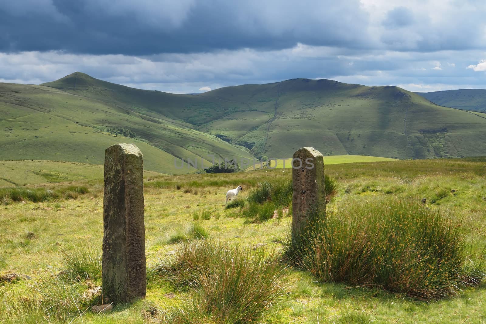 View over sunlit mountains in the Peak District National Park with old rock gate posts in the foreground and dark clouds overhead, UK