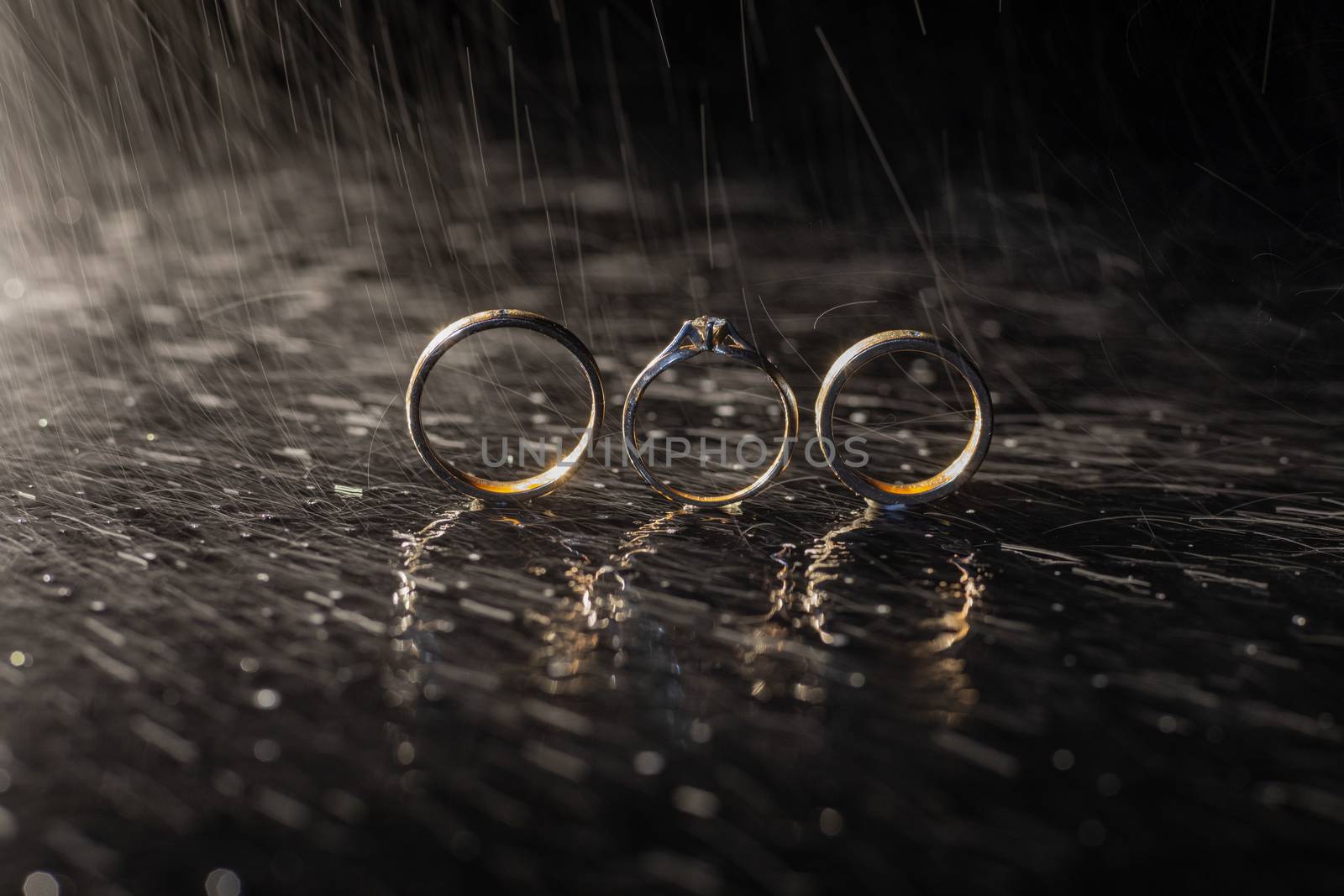 Wedding and engagement rings lying on dark surface shining with light close up macro. Water splashes. Rain