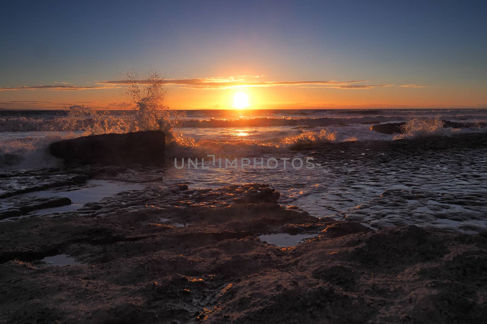 Stunning sunset with orange clouds and spray in the air from the waves crashing onto the limestone pavement of rocks on the beach at Dunraven Bay, Vale of Glamorgan, South Wales
