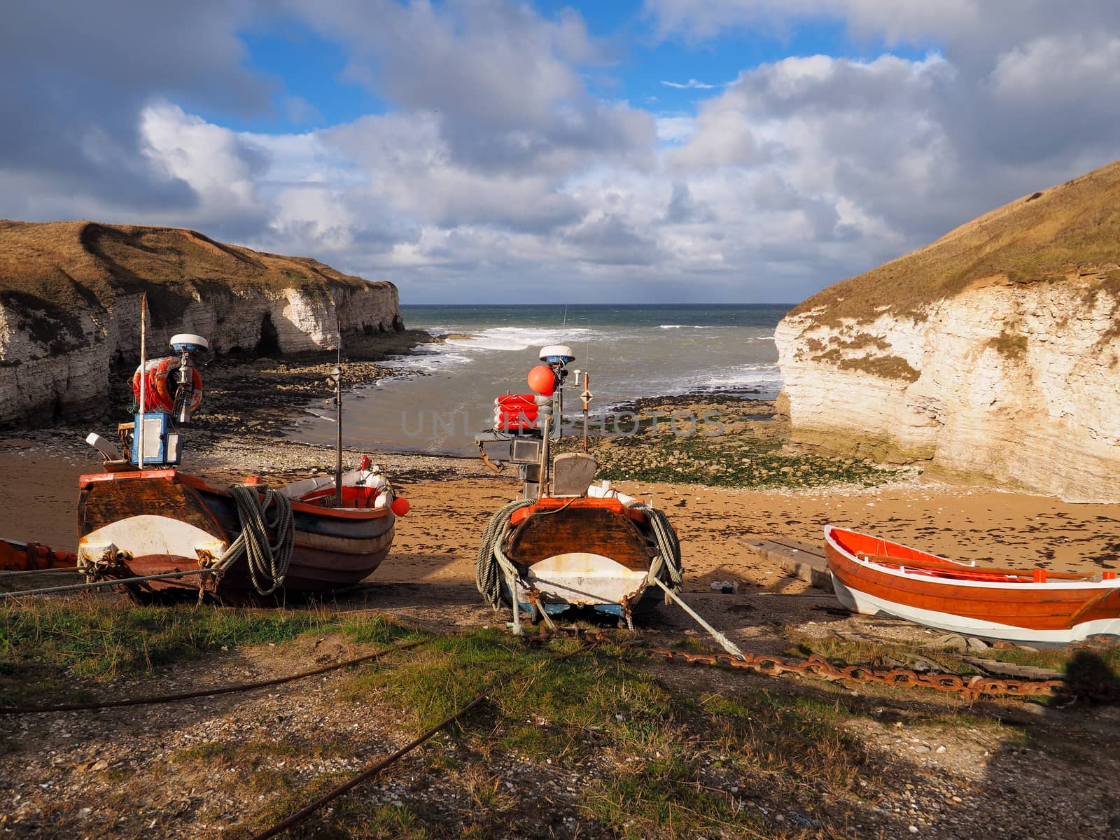 Colorful wooden and red and white fishing boats tied up on the beach in a cove waiting for the tide to come in with white cliffs in the background and a blue sky with clouds overhead.