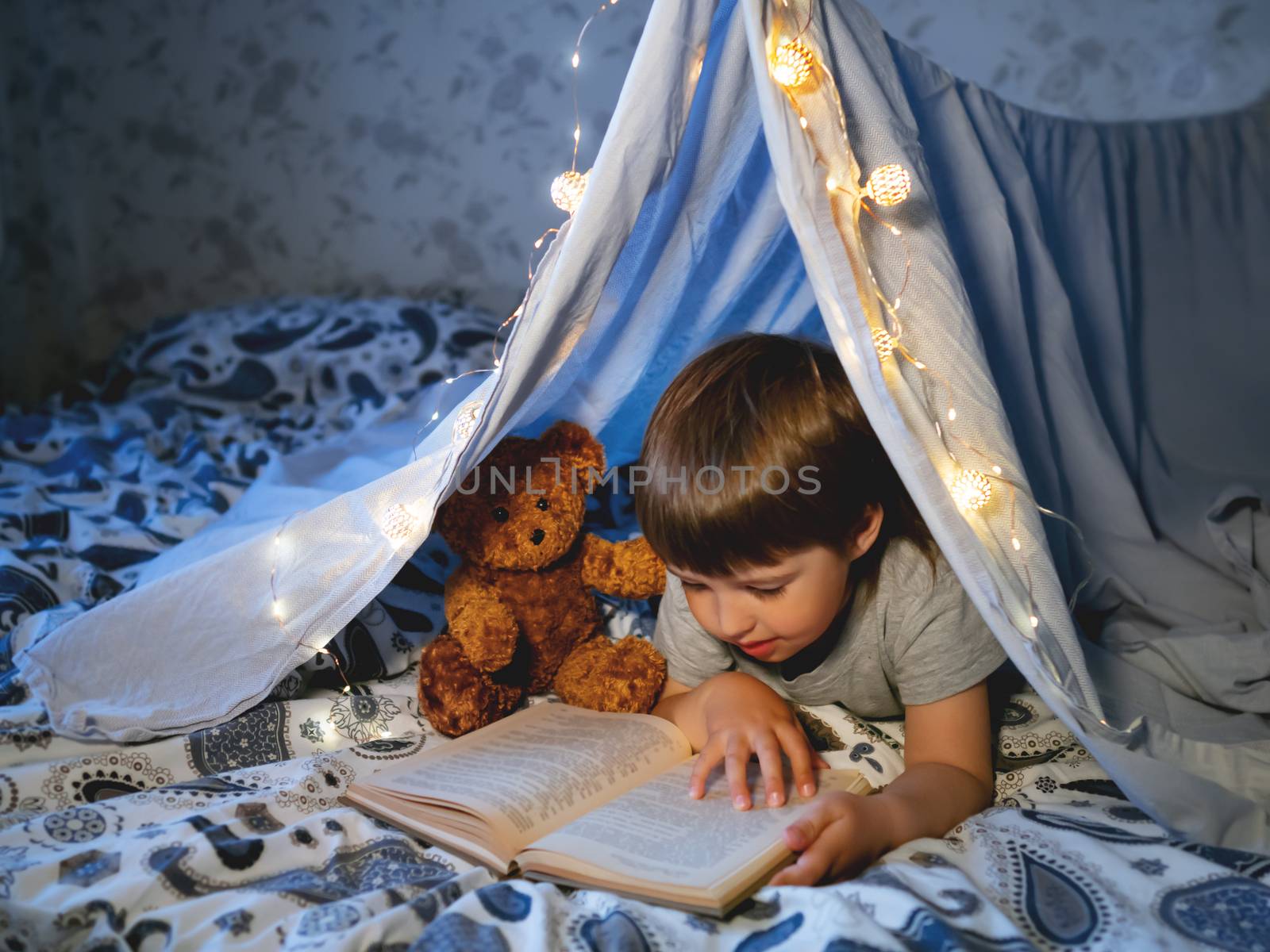 Little boy reads book. Toddler plays in tent made of linen sheet on bed. Cozy evening with favorite book.