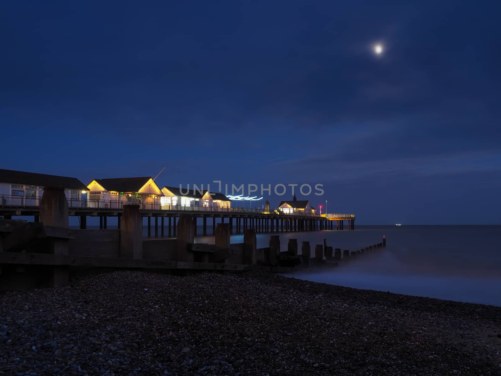 Southwold Pier with buildings and lights lit up at night and waves flowing onto a pebble beach next to a wooden groyne under a bright moon with a ship on the horizon, Suffolk, UK