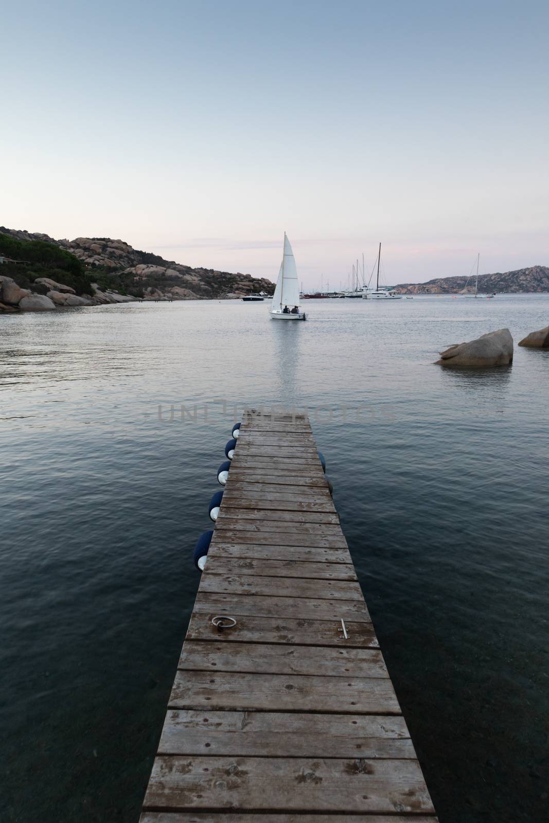 Wooden pier and sailboats sailing in evening calm sea of marvellous Porto Rafael, Costa Smeralda, Sardinia, Italy. Symbol for relaxation, wealth, leisure activity.