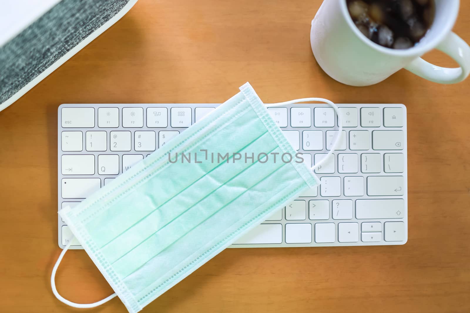 Business, Work from home, Healthcare and Coronavirus (COVID-19) Concept. Top view of surgical face mask on white computer keyboard with mug cup of iced coffee and speaker on wooden table.