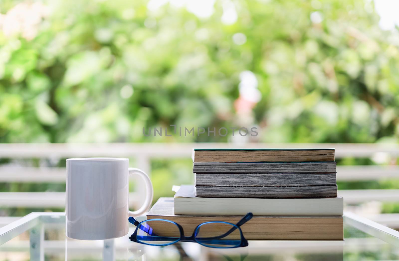 Education and relaxation Concept. Closeup of white  mug cup of hot coffee, reading glasses and books on glass table in green garden view