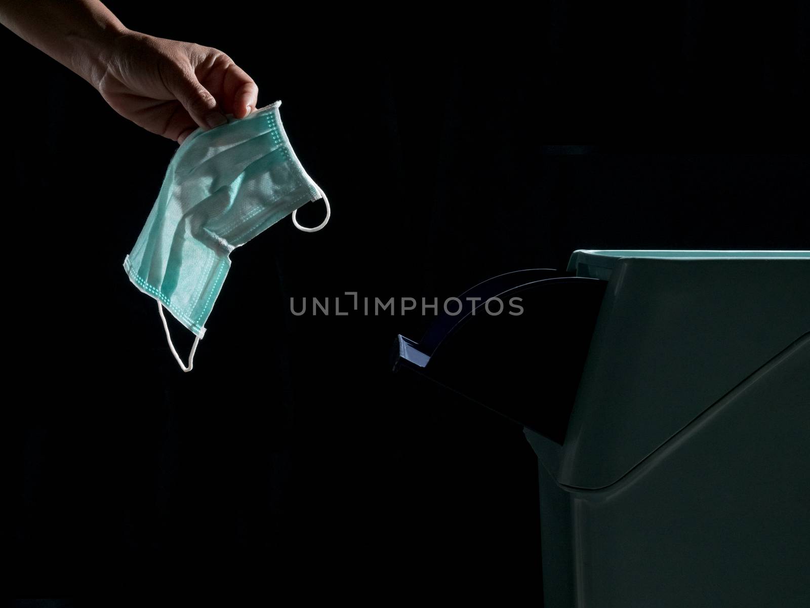 Hand putting used surgical mask to green trash, dark tone. Discarded mask concept.