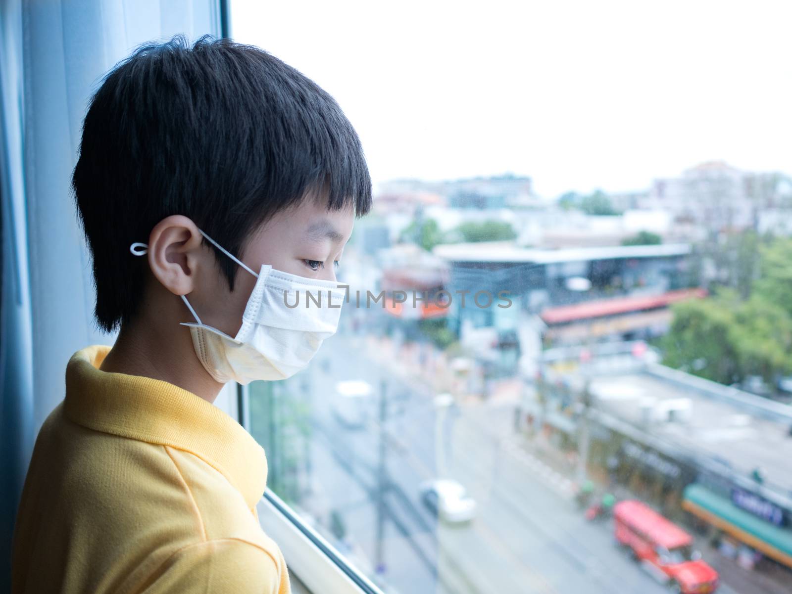 Cute asian young boy with yellow shirt,  wearing medical mask, standing idle in front of close window, looking through window, view from high building. Feel bored , long stay at home concept.